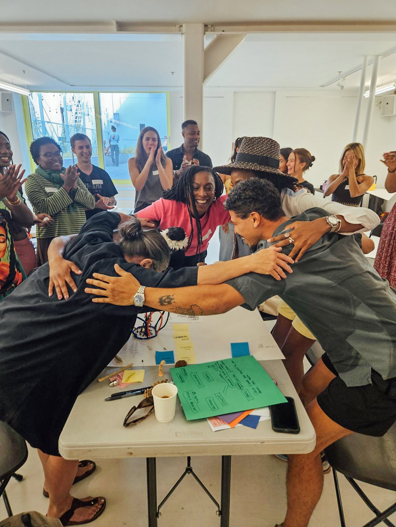 Groupe de personnes en train de se faire un câlin collectif autour d'une table, en arrière-plan, un groupe de personnes qui applaudissent et sourient dans une pièce bien éclairée.