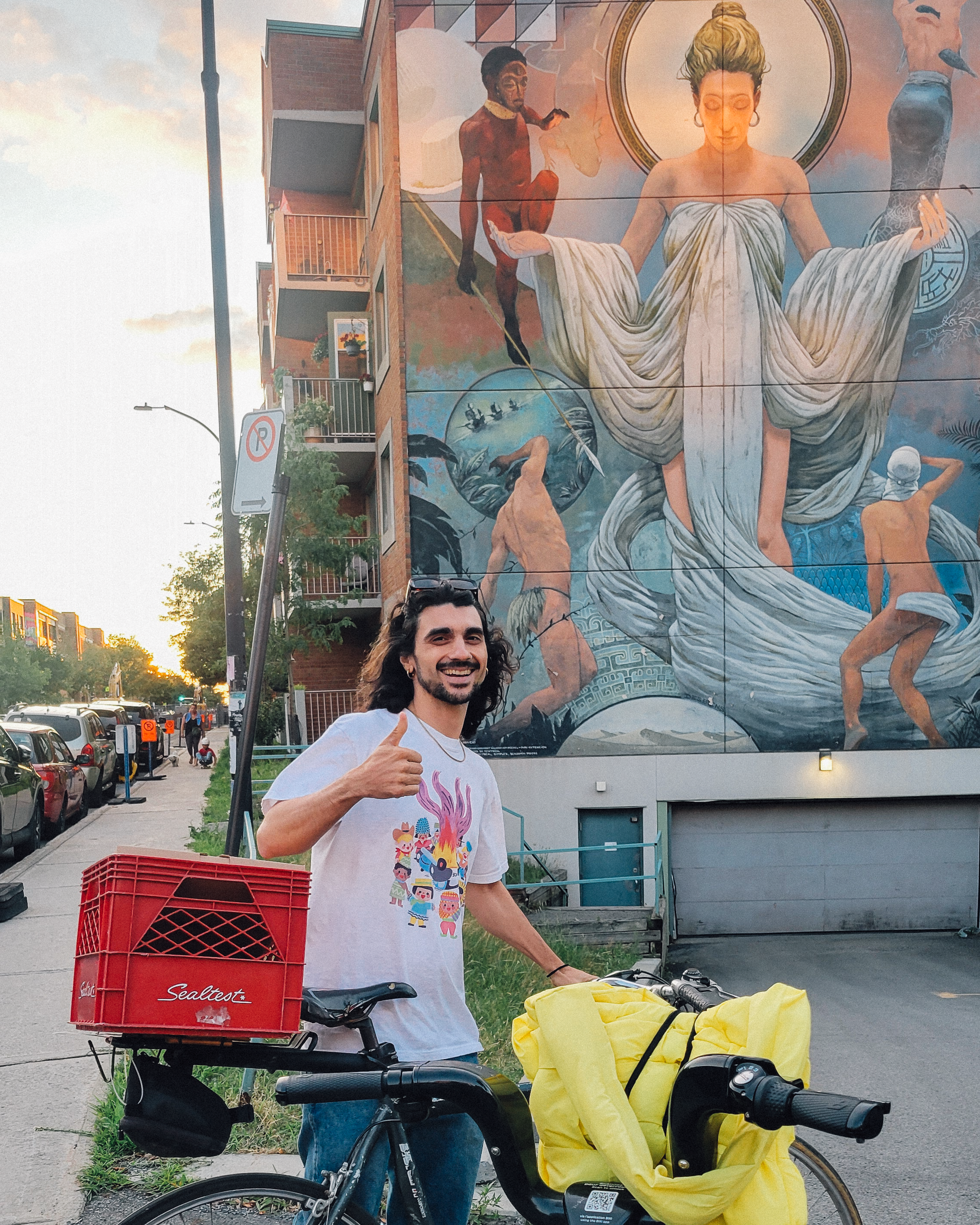Un homme souriant donne un pouce en l'air, près de son vélo équipé d'une boîte rouge Sealtex et d'une sacoche noire, avec un panneau mural représentant une peinture. Le ciel est clair et des bâtiments résidentiels sont visibles en arrière-plan.