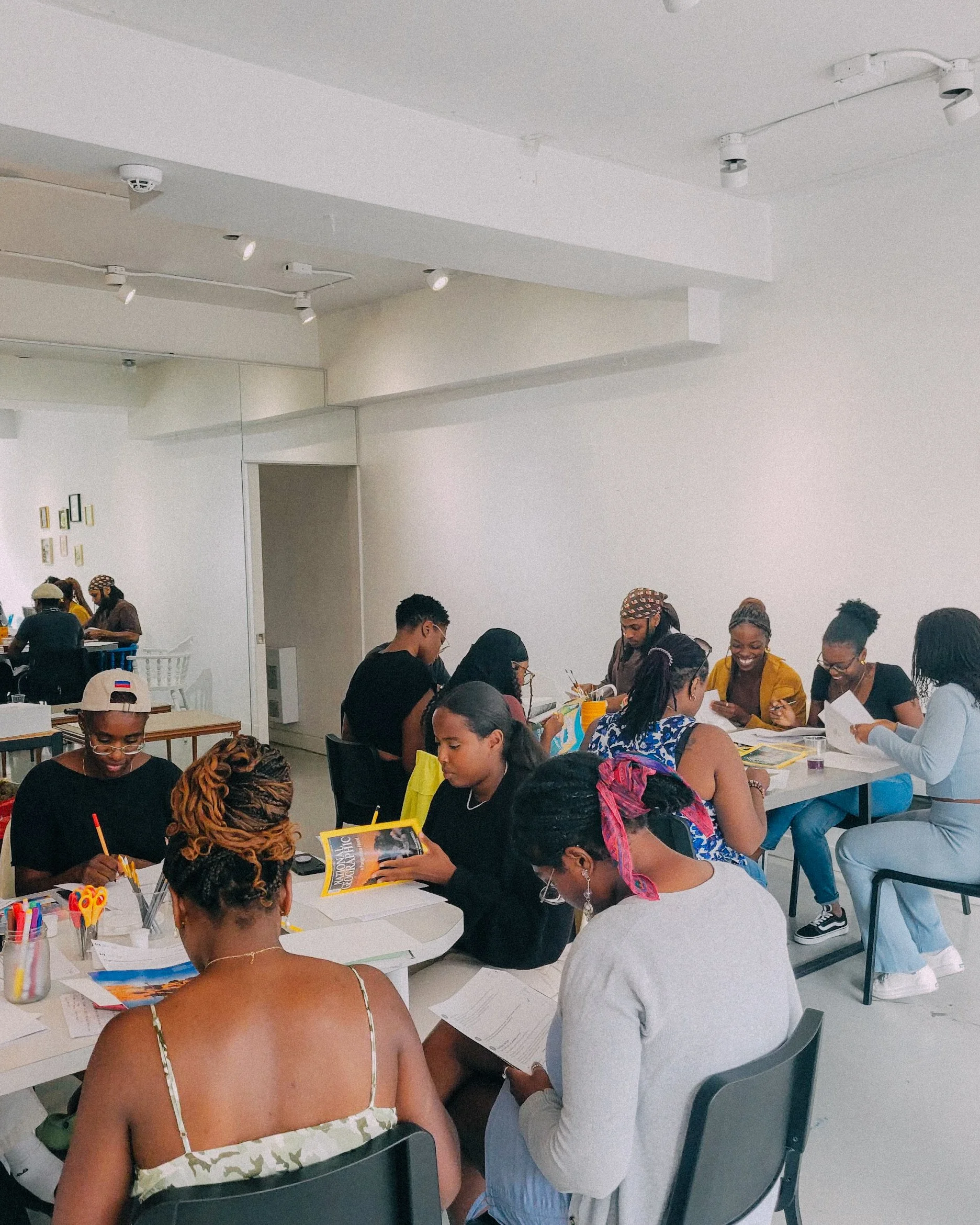 Un groupe de personnes, majoritairement de couleur, assises autour de tables dans une pièce blanche, en train de lire, écrire ou discuter, avec des fournitures scolaires ou artistiques sur la table.