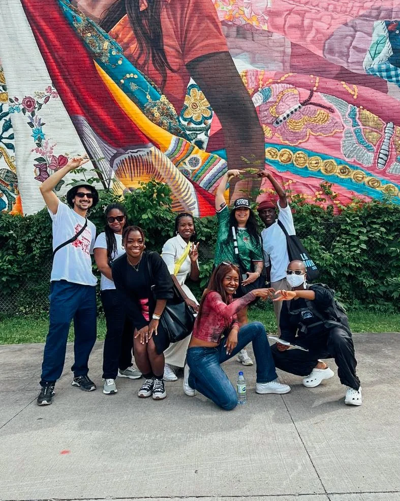 Groupe de huit personnes posant devant une fresque colorée représentant des femmes en vêtements traditionnels en rouge avec un fond de verdure.