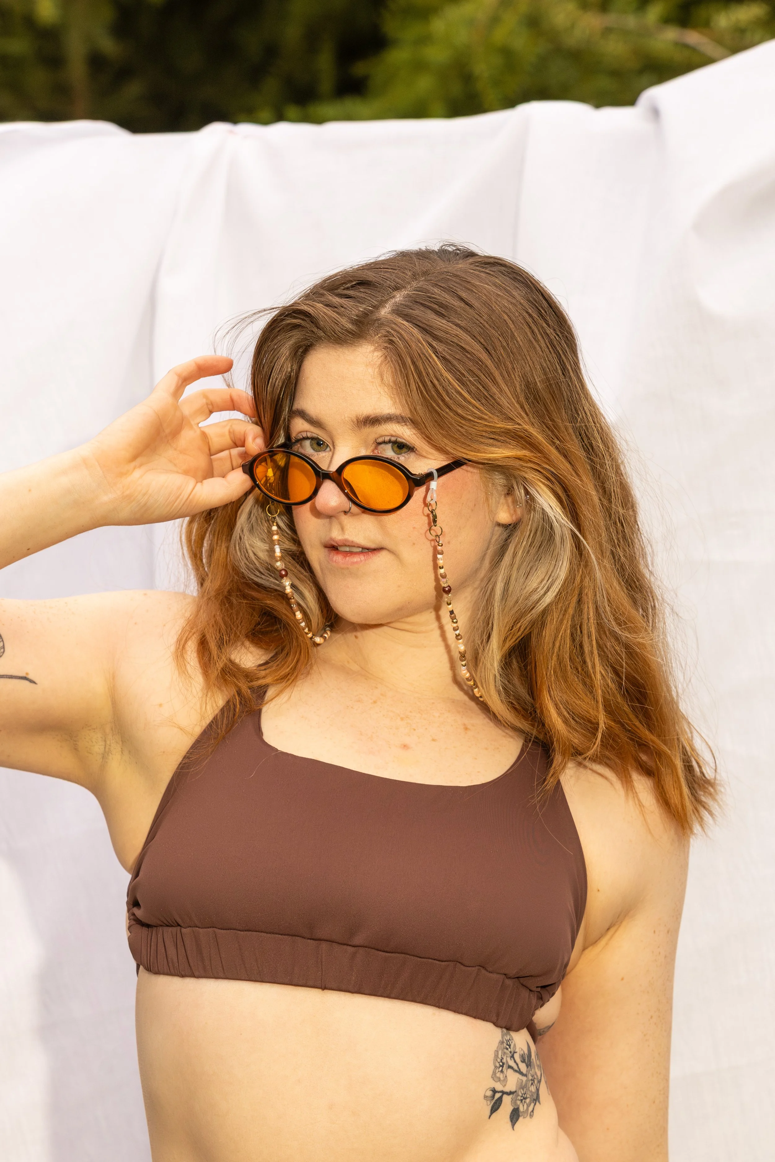 A woman with red hair and light skin is adjusting orange-tinted sunglasses with her left hand. She is wearing a brown sleeveless top and has a tattoo of flowers on her left side near her waist. Behind her is a white backdrop and some green trees in the background.