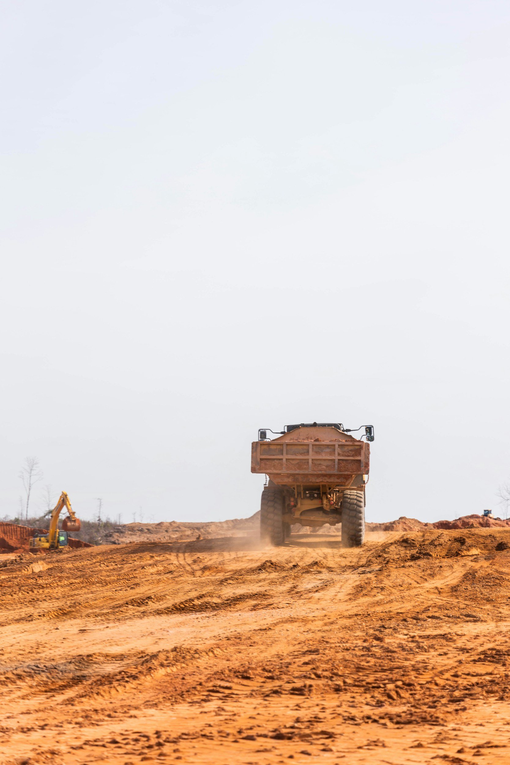 A large construction vehicle, possibly a dump truck, driving on a dirt construction site with smaller excavators in the background and a cloudy sky above.
