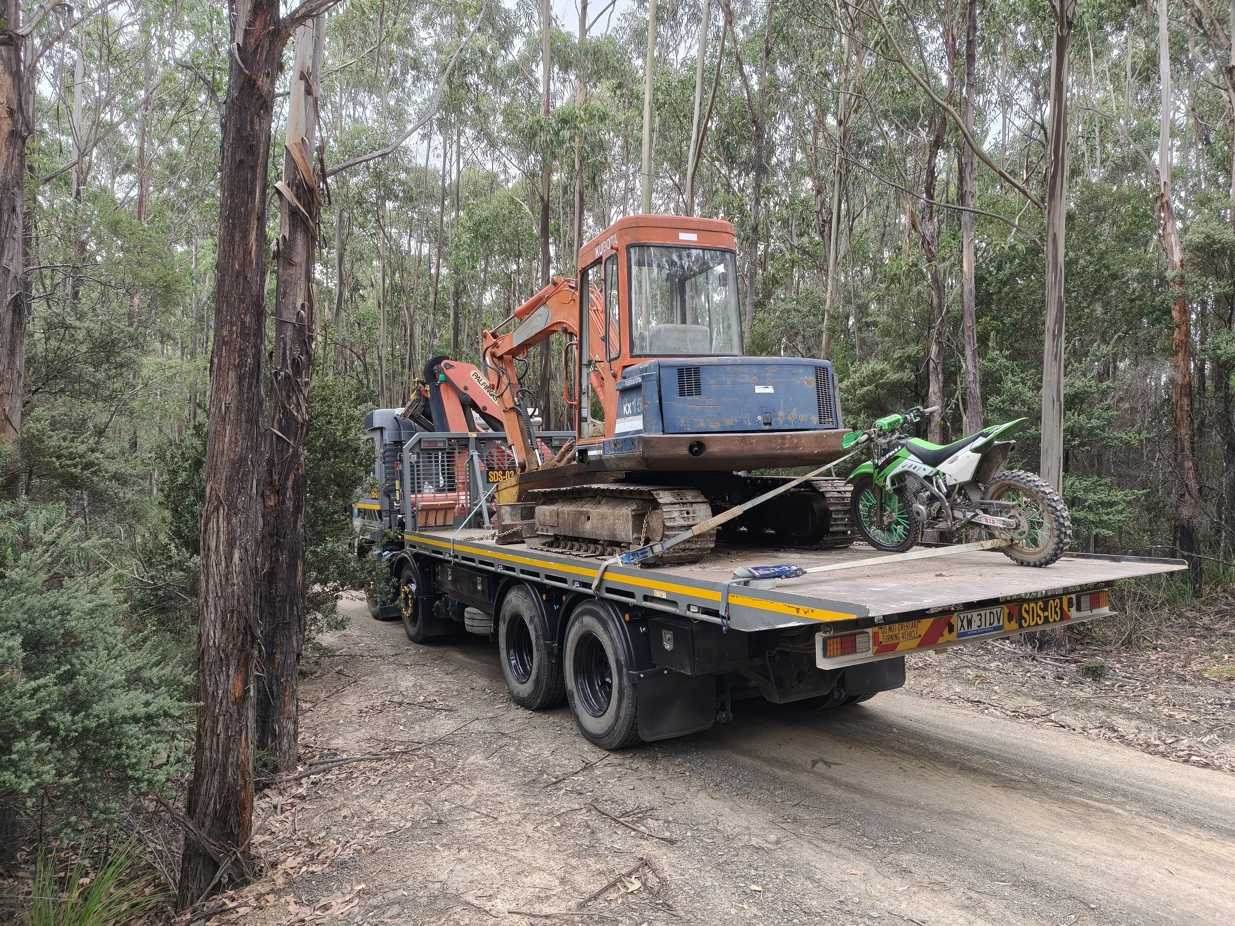 A flatbed truck in a wooded area carrying construction equipment, including an orange and blue excavator, with a green dirt bike secured alongside.