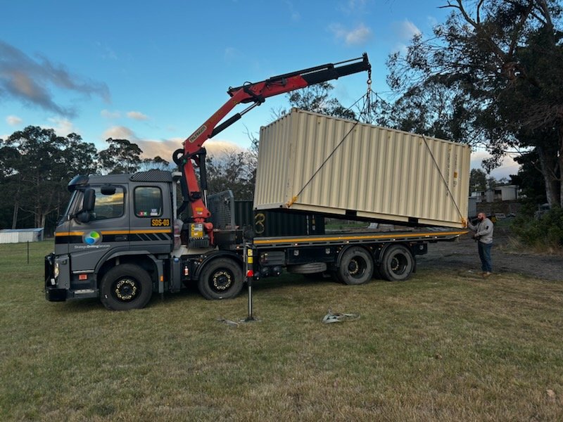 A flatbed truck with a mounted crane lifting a large beige shipping container into place, with a person guiding the process outdoors on grass and trees in the background.