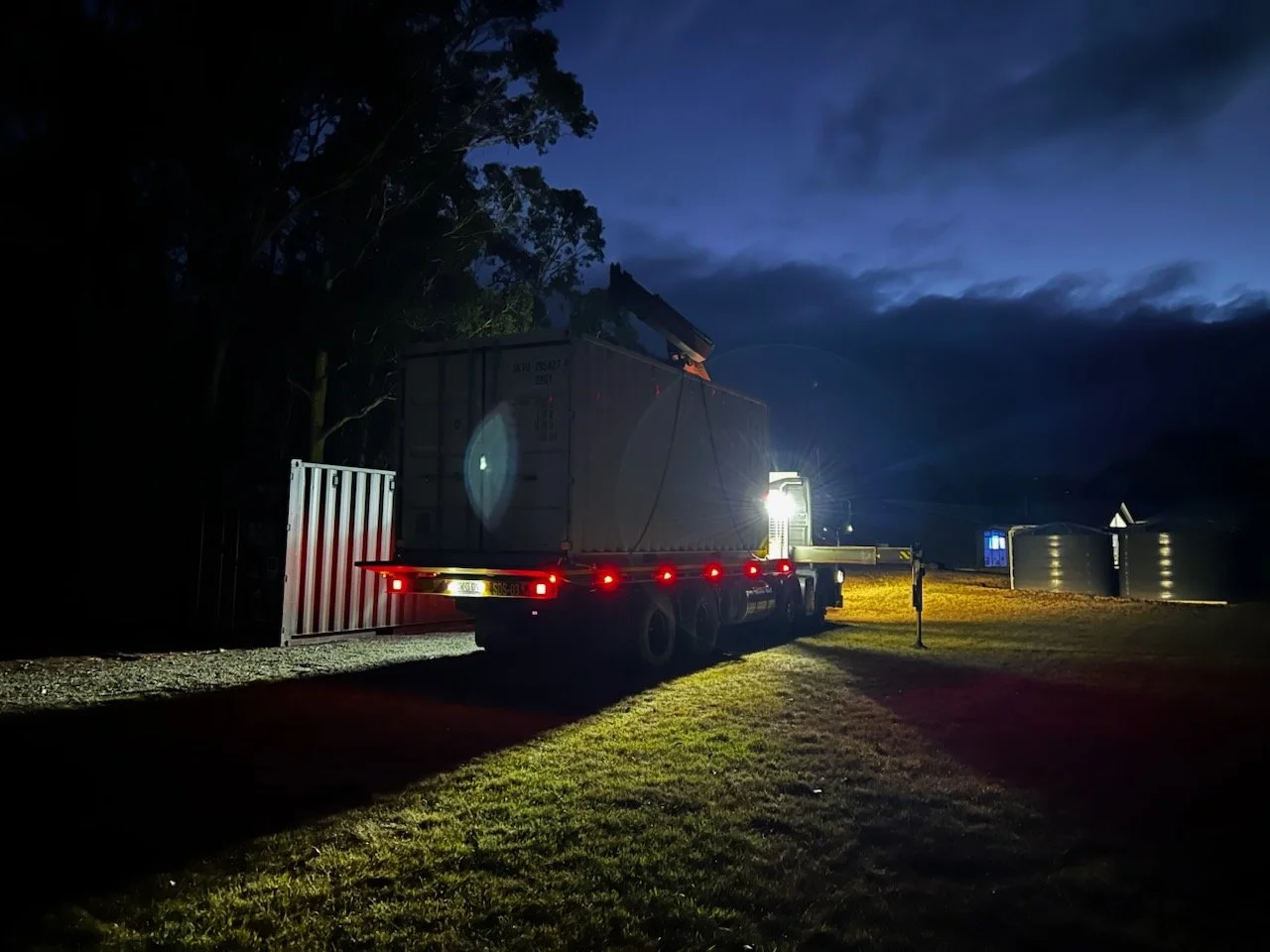 A truck carrying a large container is parked outdoors at dusk with its headlights on, illuminating the surrounding area and casting shadows on the grass. There are trees and water tanks in the background.