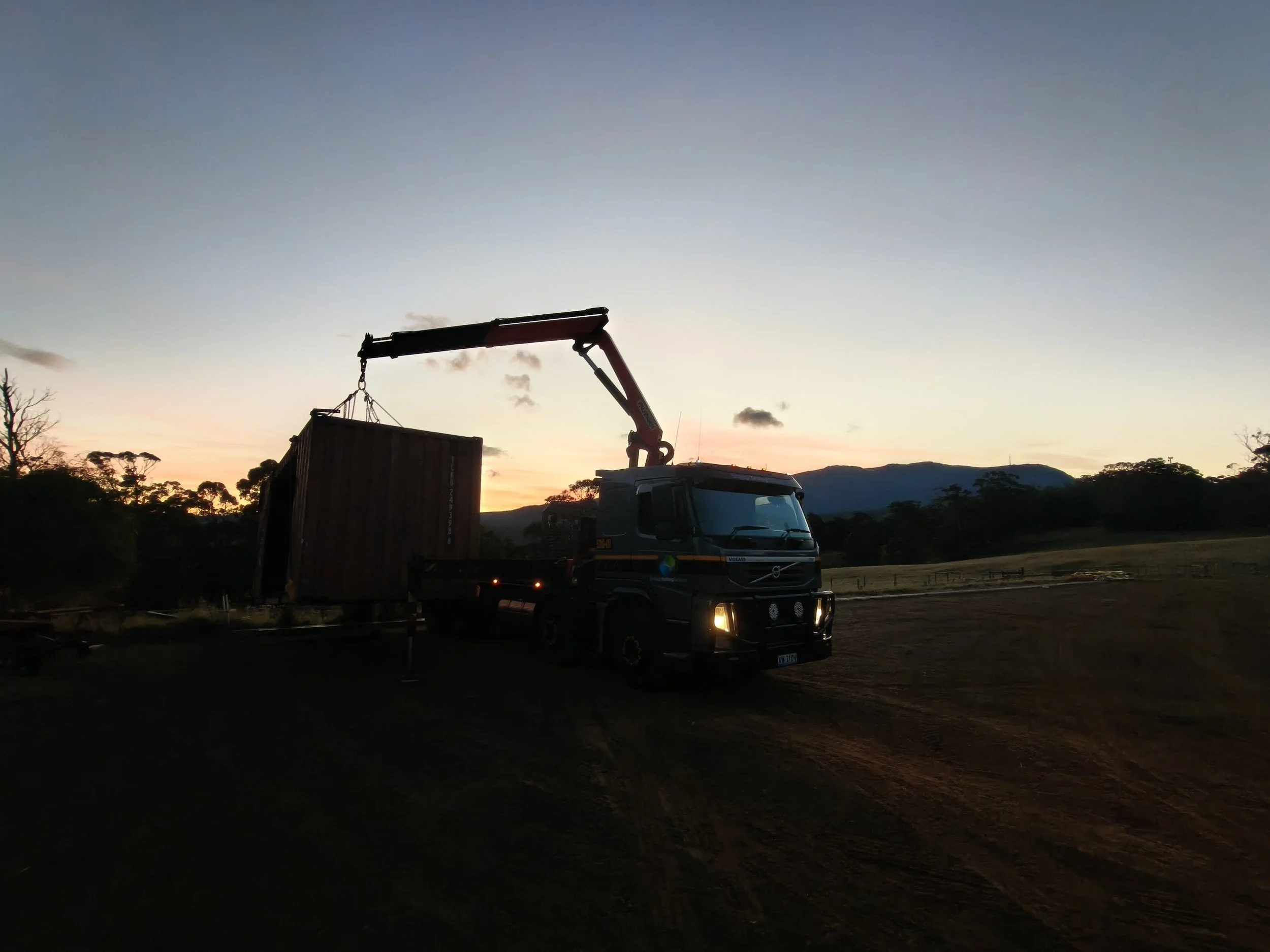 A truck with a crane lifting a container, set against a sunset sky with mountains and trees in the background.
