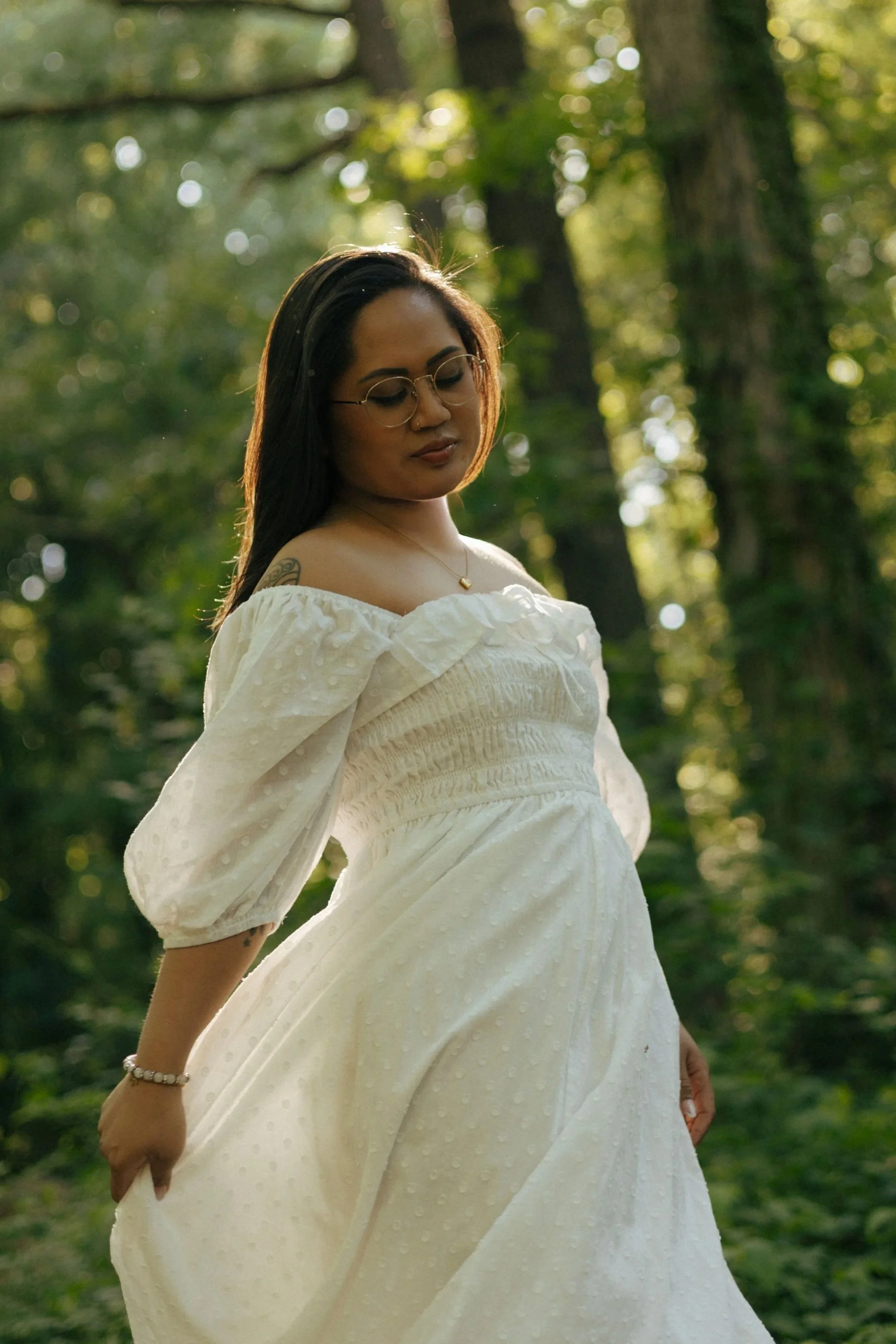 A woman standing outdoors in a forested area, wearing a white off-shoulder dress with puffed sleeves and glasses, looking down with a serene expression.