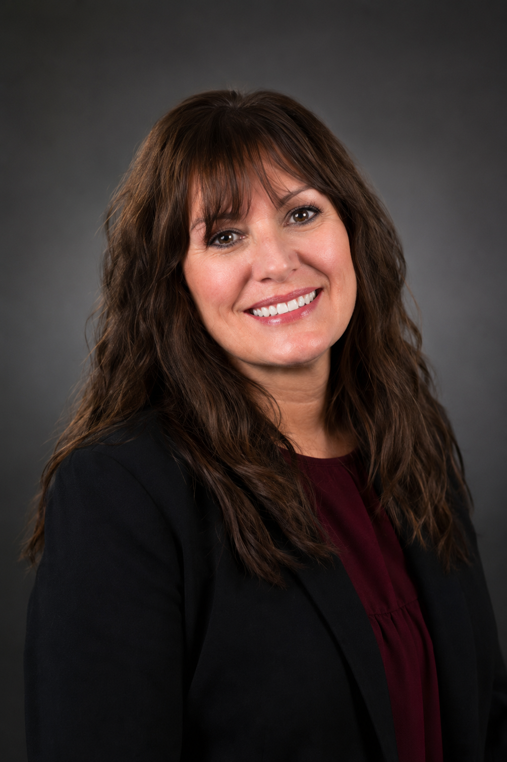 Woman with brown hair black jacket and burgundy shirt in front of grey backbround