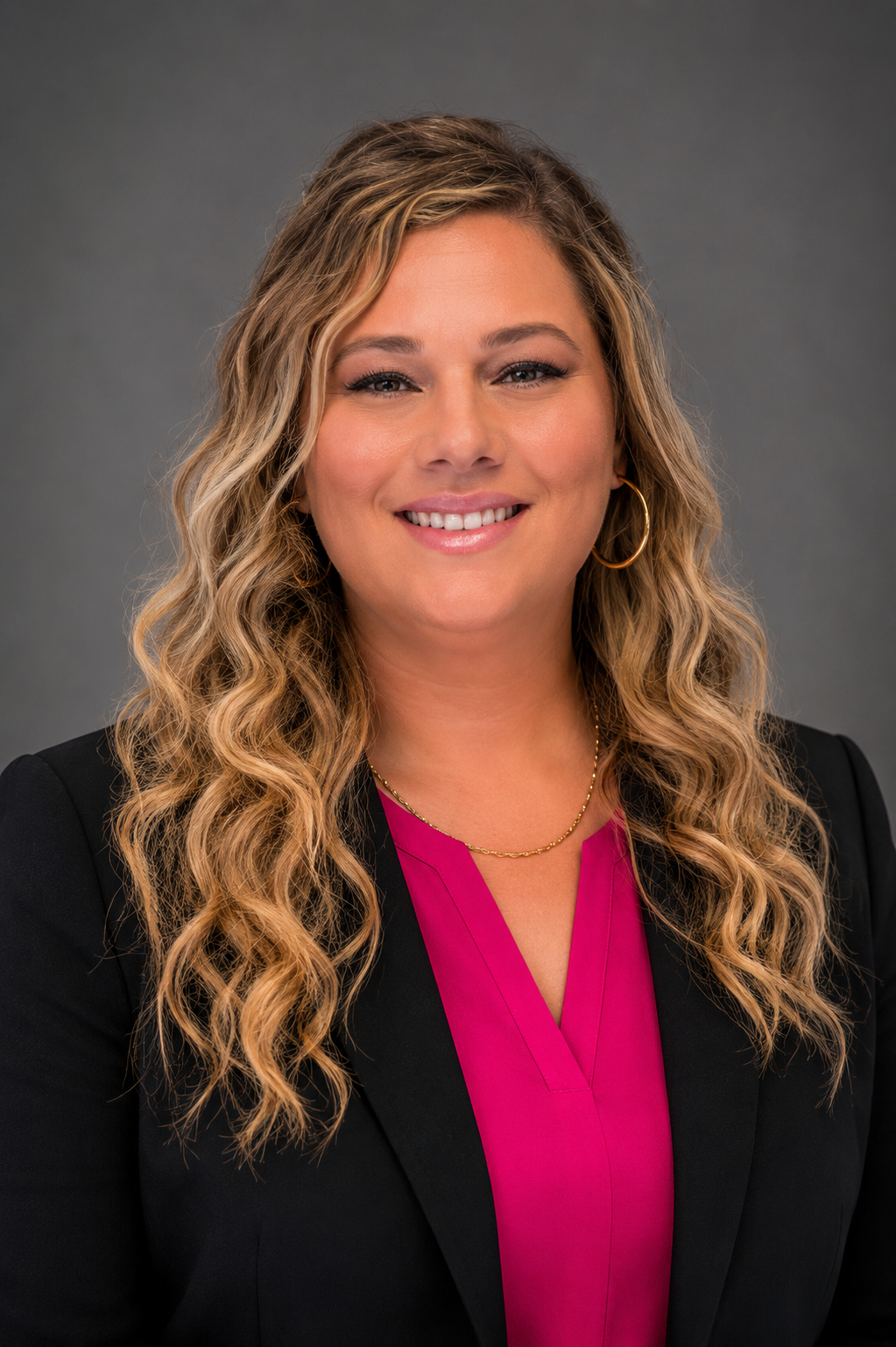 woman with pink shirt and black jacket smiling in front of grey background