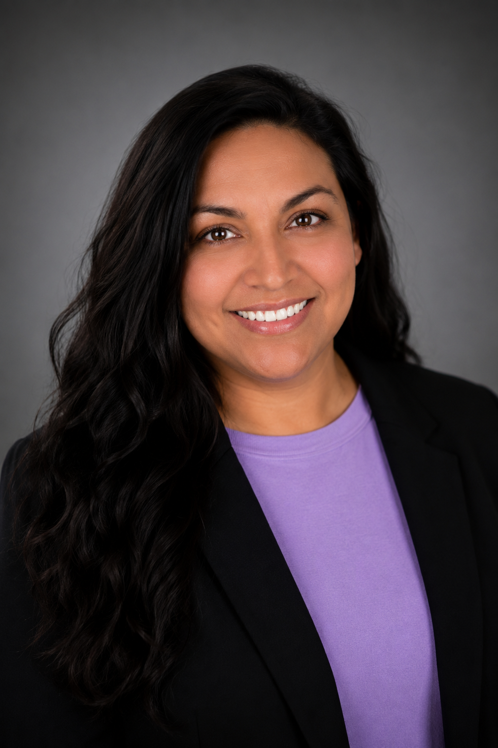 Woman with brown hair, black jacket and purple shirt in front of grey background