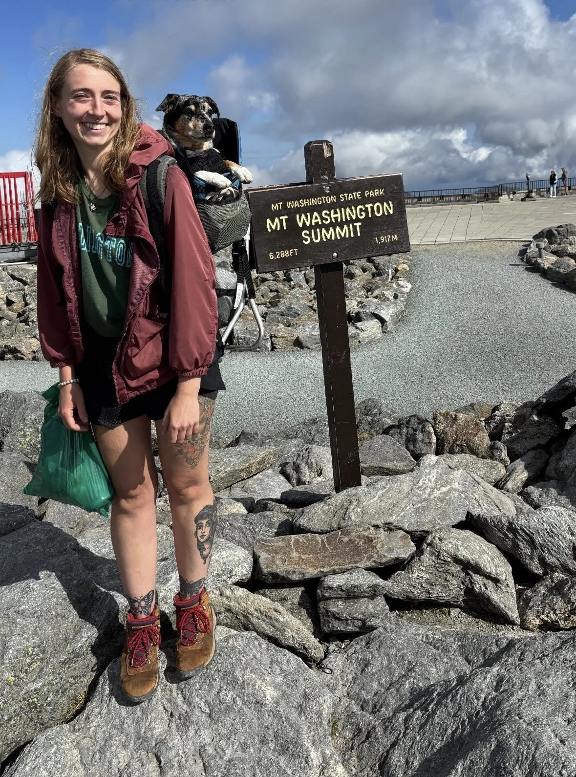 A young woman with tattoos on her legs standing on rocks at Mount Washington summit, smiling, with a dog in a backpack carrier, near a sign that reads 'Mt Washington, 6,288 ft, 1,917 m.' The sky is partly cloudy with some blue showing.