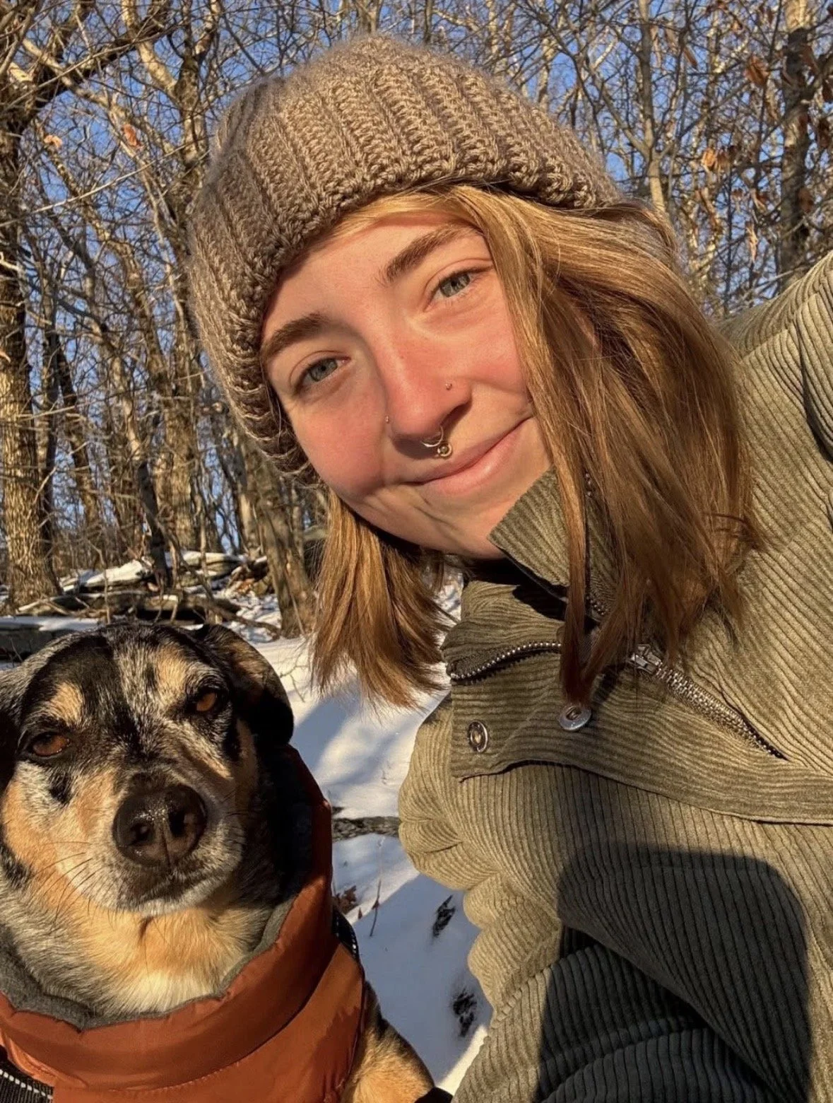 A young woman with shoulder-length reddish hair, wearing a brown knit beanie and a beige jacket, smiling and taking a selfie outdoors in a snowy forest with a dog beside her. The dog is wearing an orange jacket, has a black and tan coat, and is sitting on the snow.
