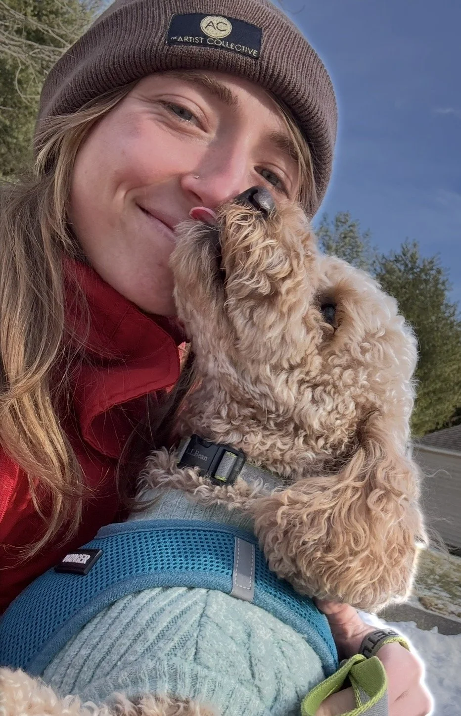 A woman in a brown beanie hat smiles as her dog licks her face outdoors on a sunny day. The woman is wearing a red jacket and the dog is a curly-haired breed, wearing a harness, with a blue and green backpack nearby.