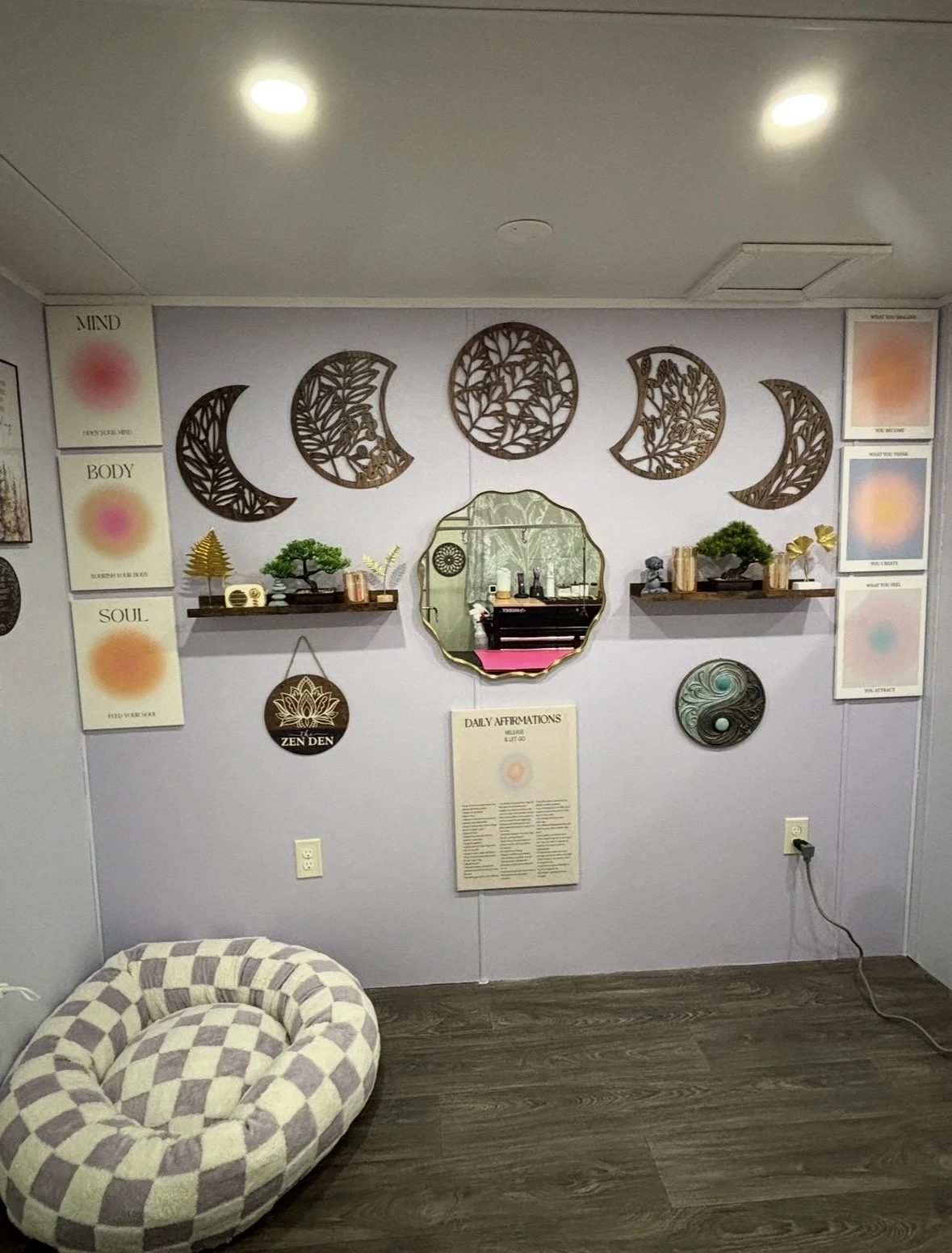 A wellness studio with wall decor featuring moon phases and affirmations. A mirror reflects a black piano, and a cushioned seating area with a checkered pattern is in the foreground. The wall has shelves with plants and decorative items.