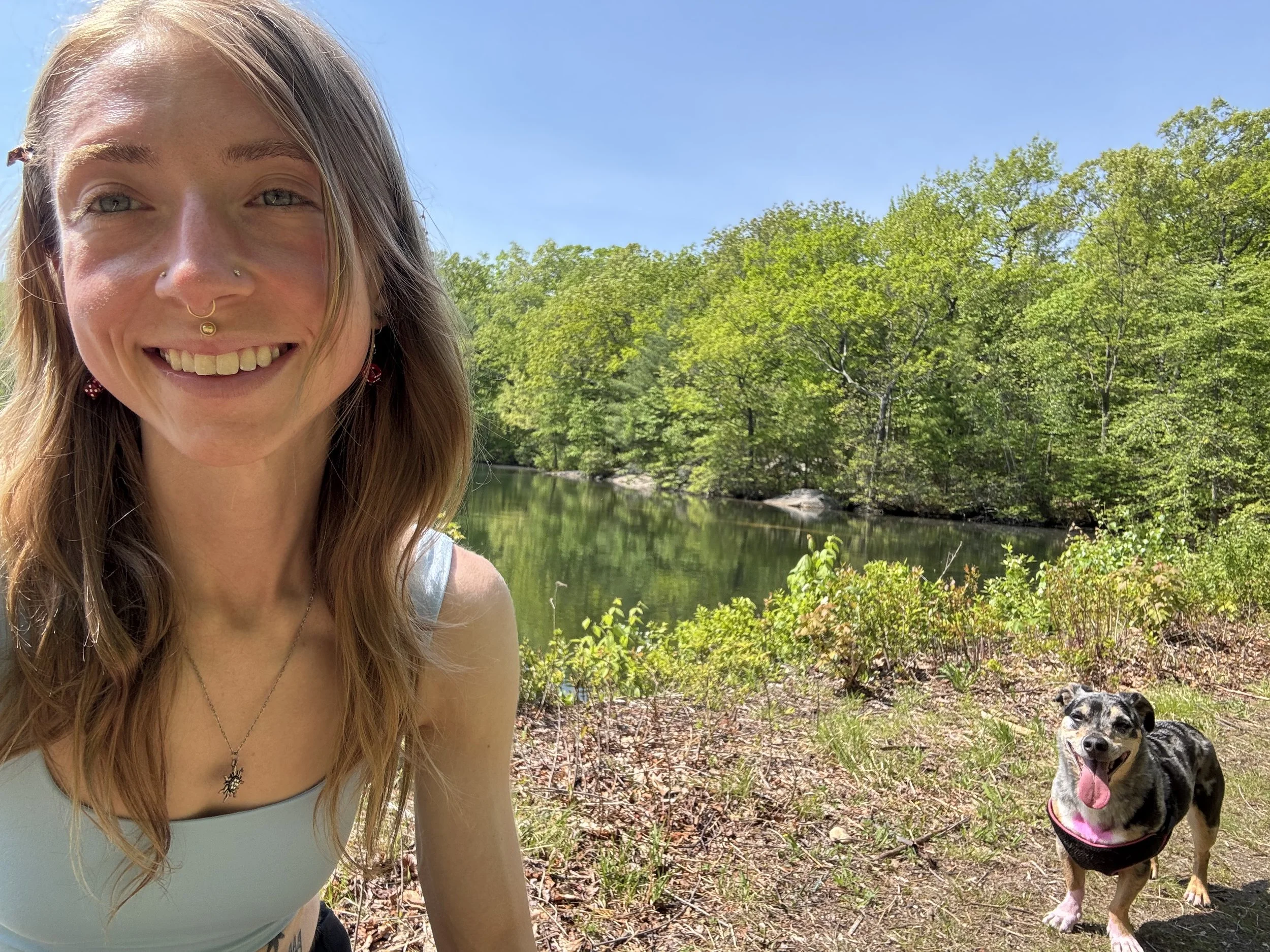 A smiling young woman with blonde hair and blue eyes takes a selfie outdoors near a river, with a black and white dog wearing a pink harness nearby.