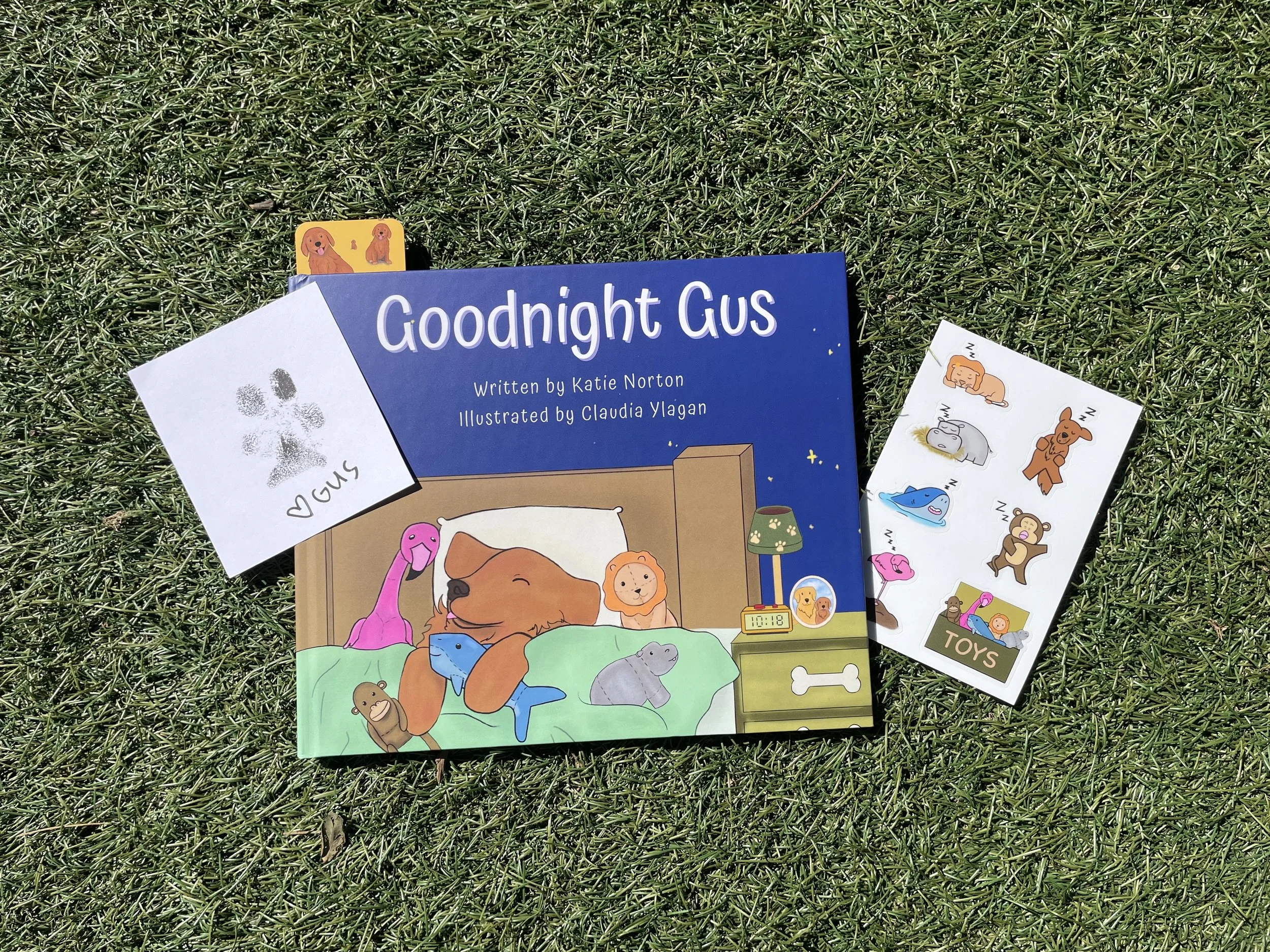 A children's book titled "Goodnight Gus" by Katie Norton, illustrated by Claudia Ylagan, is placed on grass, accompanied by various stickers and a small sticker of a dog. The book cover features illustrated animals, including a dog, giraffe, and elephant, in a cozy setting.