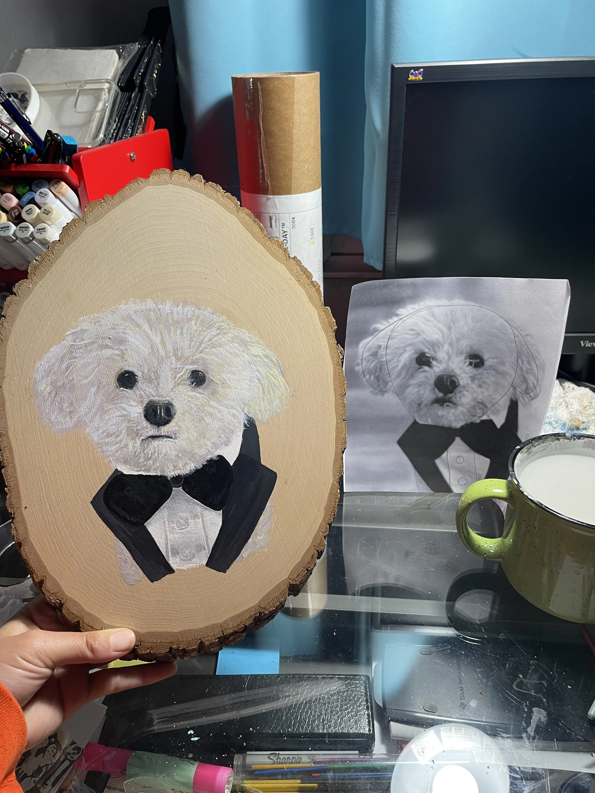 A wooden art piece of a dog dressed in a tuxedo, painted with realistic details, held by a hand. A printed black and white photo of the same dog is on the right, on a table surface with art supplies.