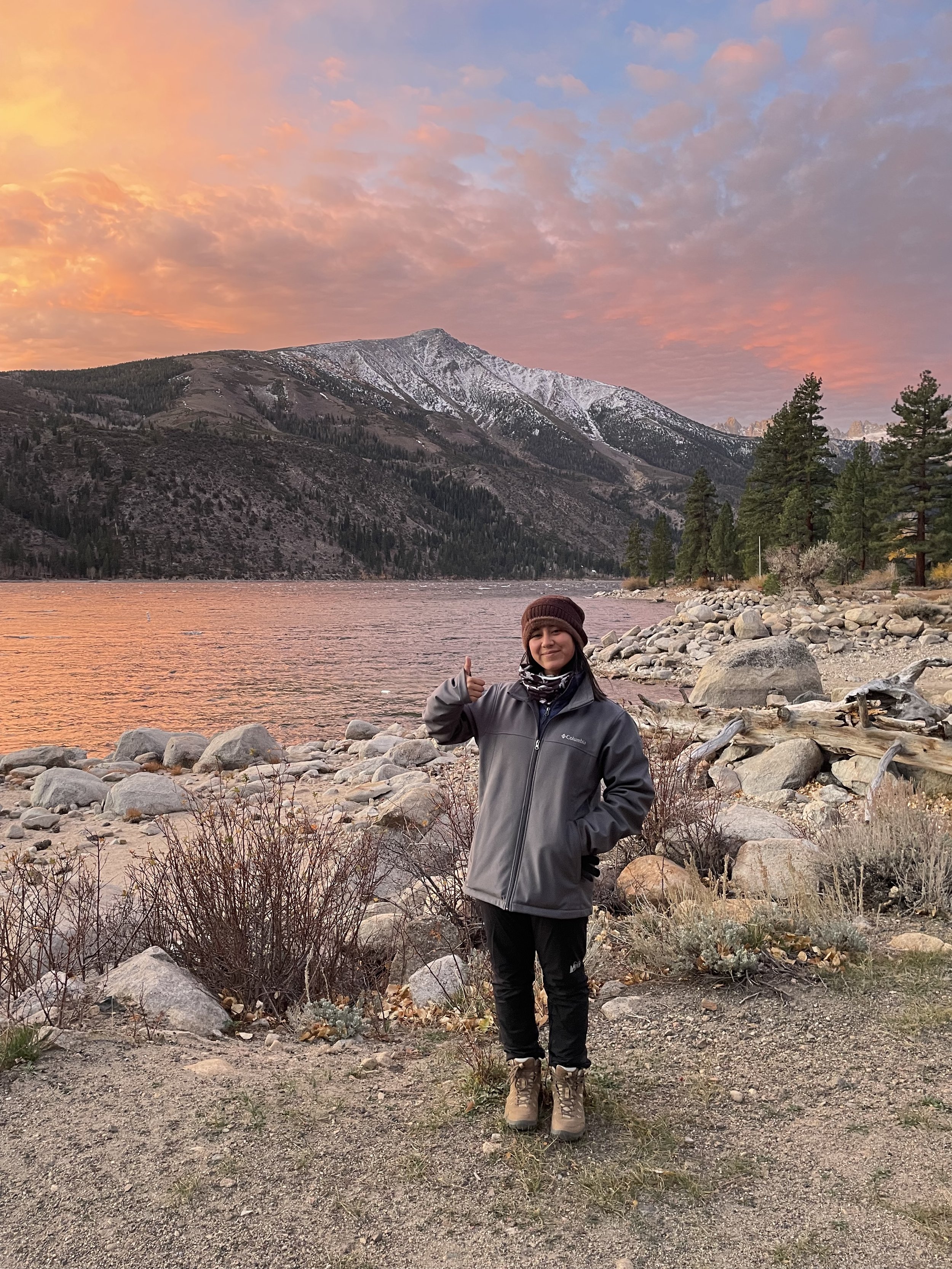 A woman in outdoor clothing standing on rocky terrain near a body of water with trees, snow-capped mountains, and pink and orange clouds in the background during sunset.