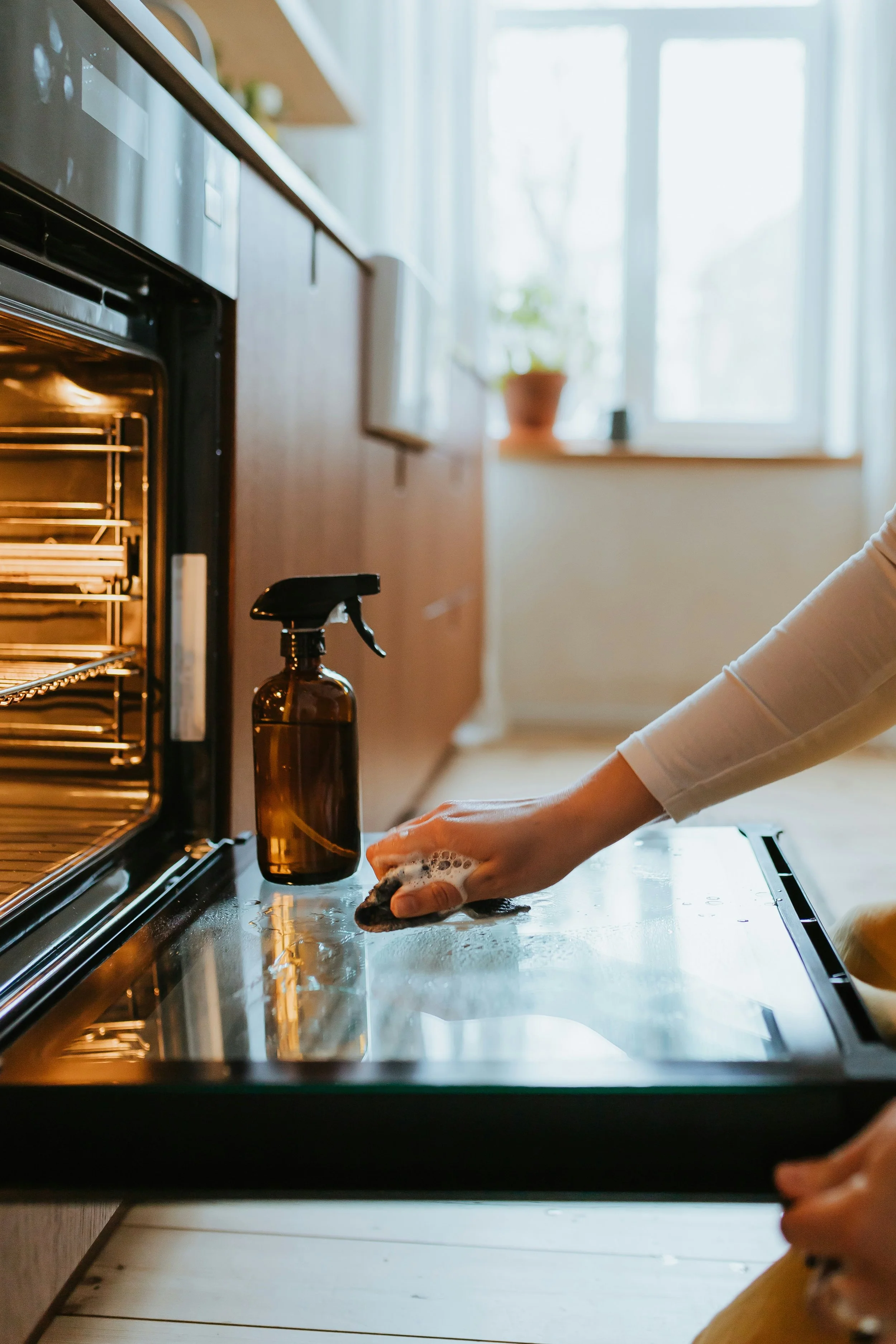 A person cleaning the kitchen countertop with a sponge, with a spray bottle nearby, in front of an oven, in a bright room with a window and a plant in a pot in the background.