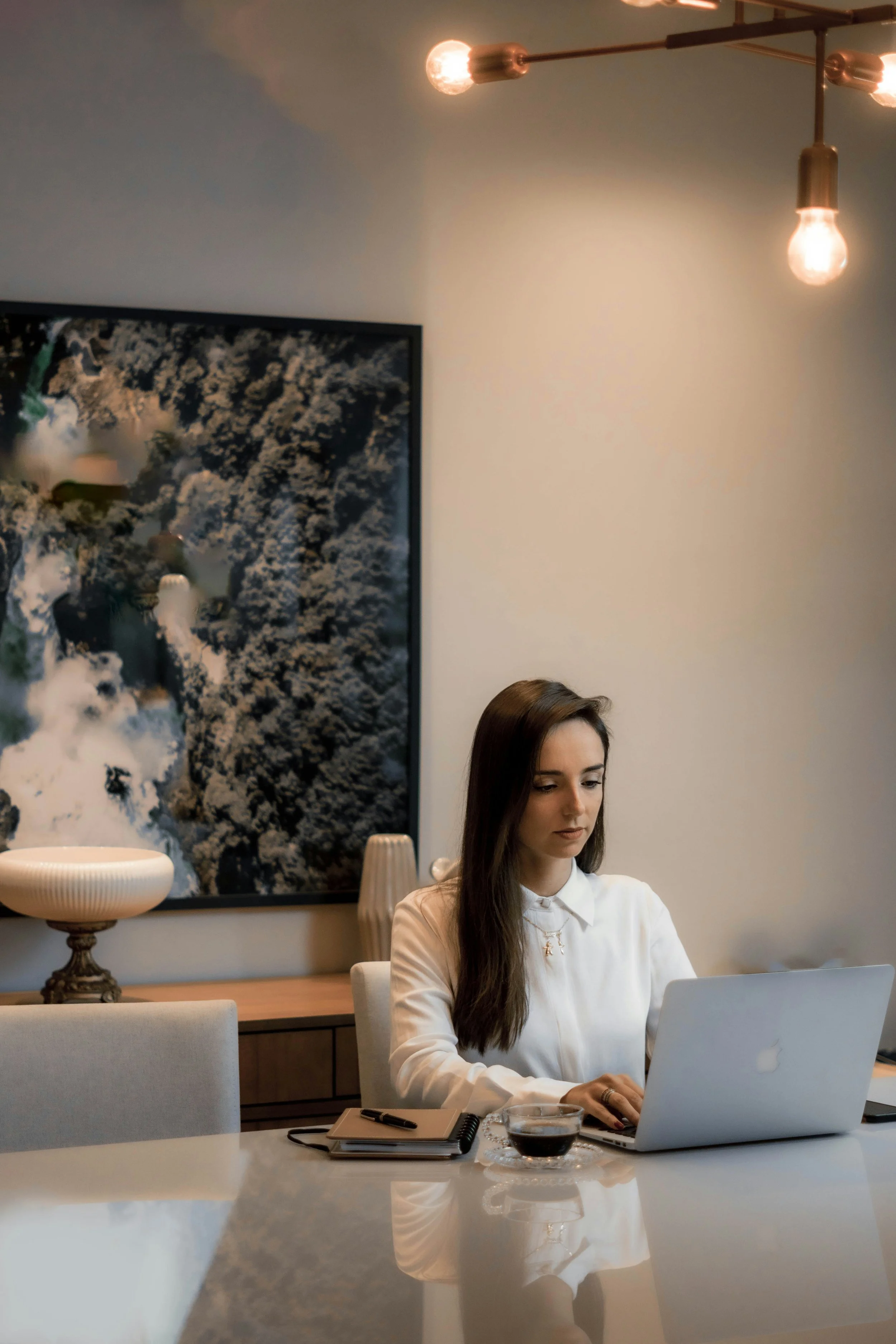 A woman with long brown hair wearing a white blouse, sitting at a table working on a laptop in a modern, well-lit room with contemporary decor. A large, abstract black and white artwork hangs on the wall behind her, and there are decorative items like a bowl and a cup on the table.