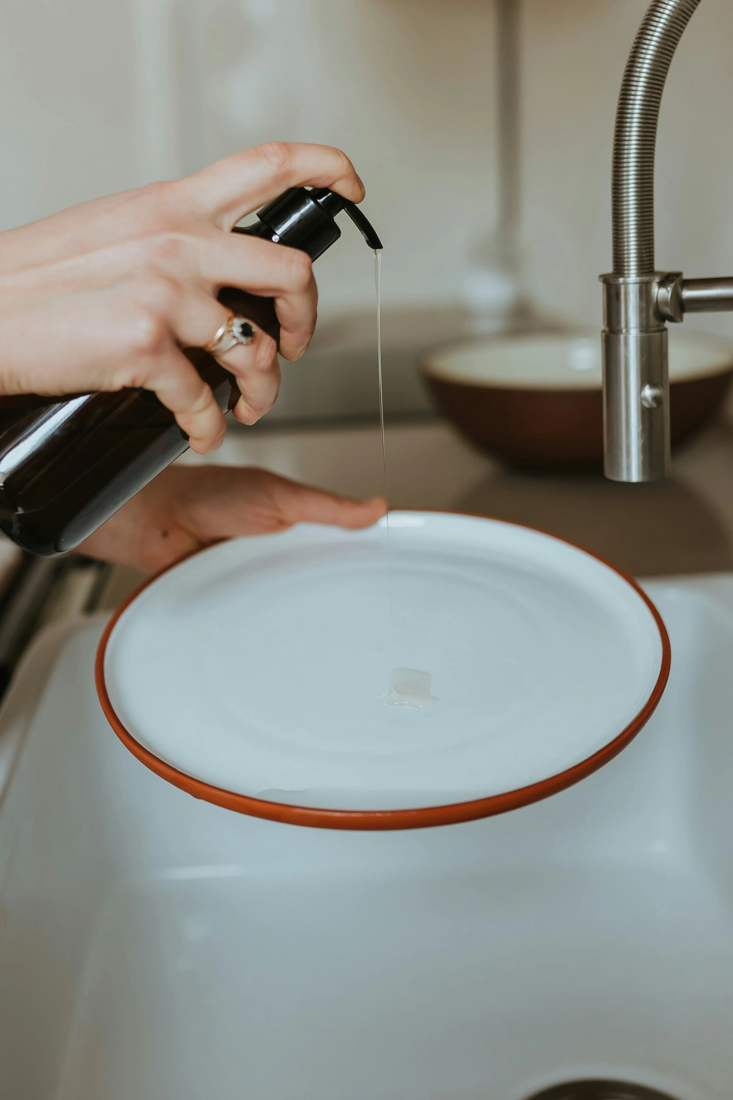 Close-up of a person pressing a black soap dispenser with their right hand, water flowing into a white bowl with a red rim underneath a kitchen faucet.
