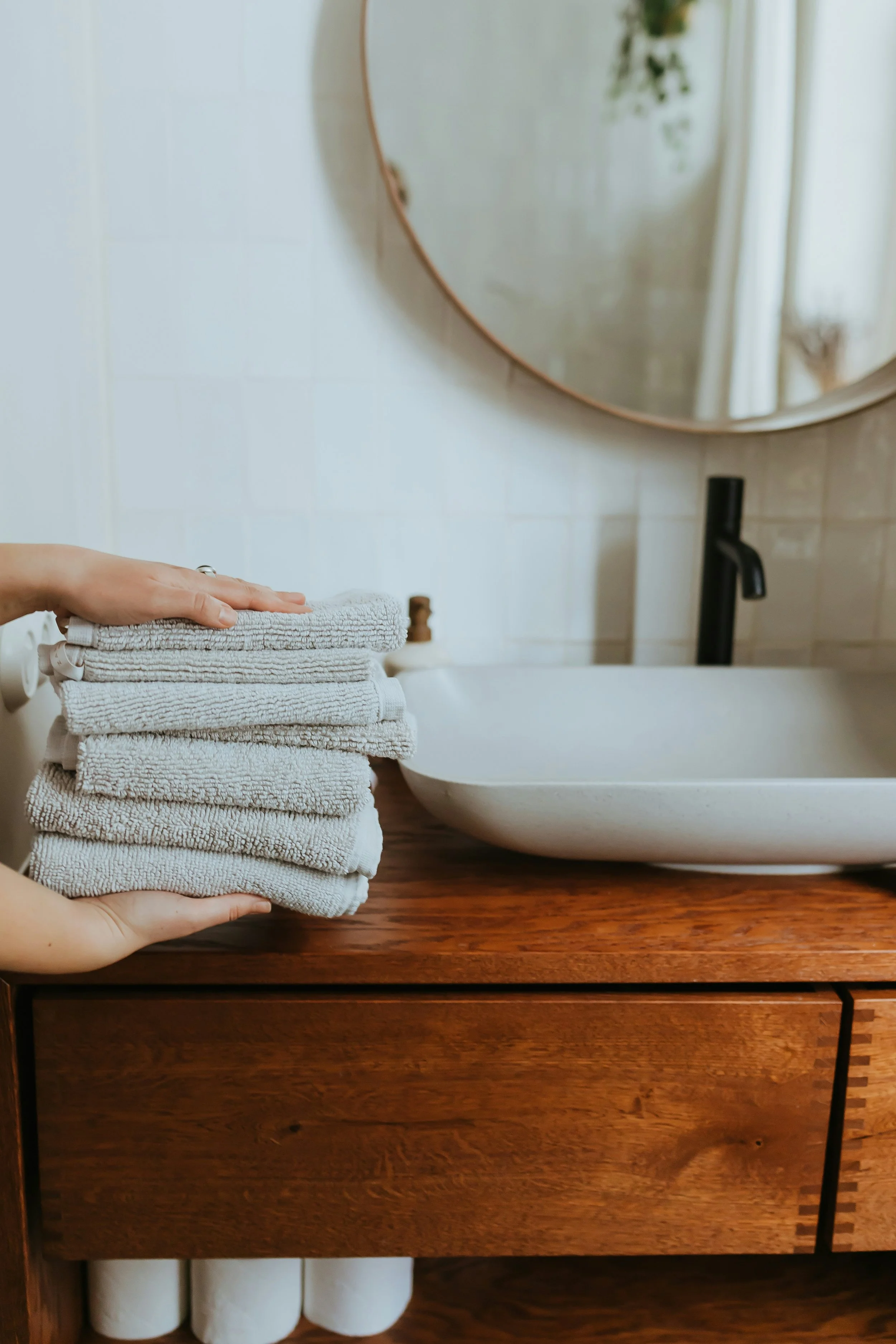 Person holding folded gray towels on a wooden counter, bathroom with a round mirror and white tiled wall in the background.