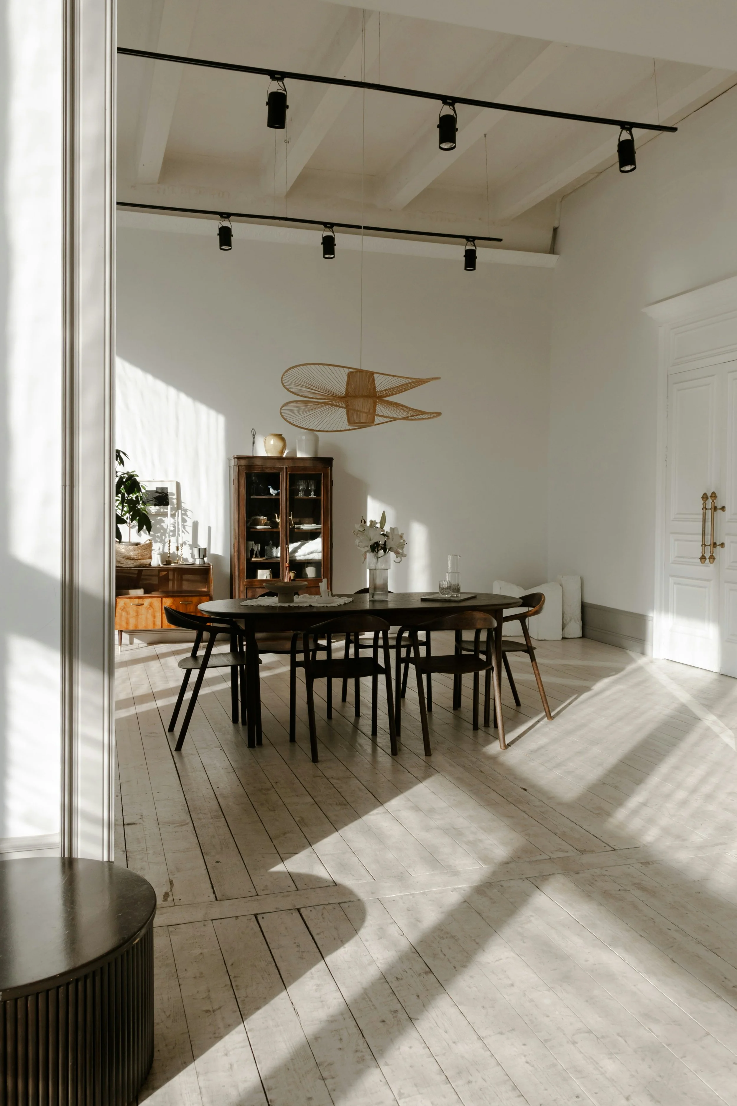 A dining room with white walls, a wooden floor, a black dining table with chairs, and a wooden cabinet. There is a modern hanging light fixture and a ceiling with exposed beams. Natural light creates shadows across the room.
