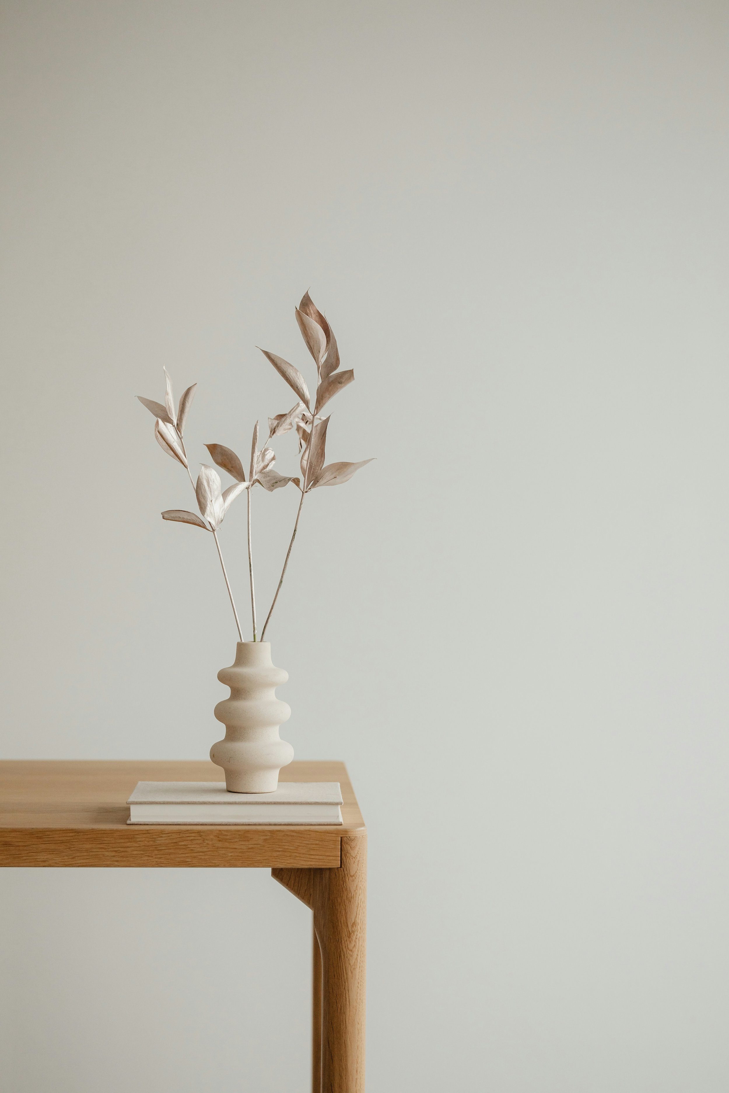 A beige vase with a unique shape and horizontal ridges, holding dried beige leaves, placed on a wooden table with a light-colored cloth underneath, against a pale wall.