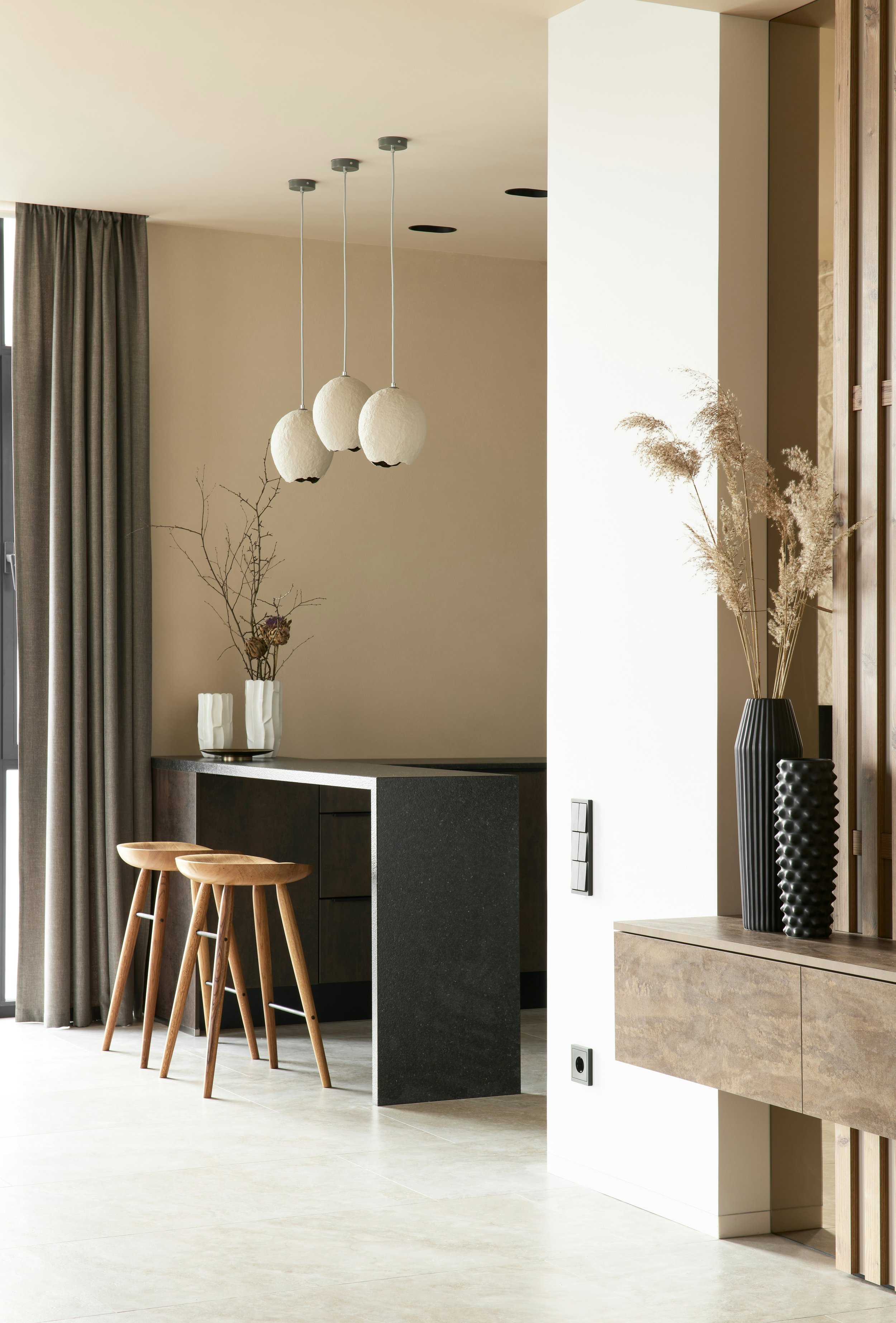 Modern interior with a black kitchen island, light wood bar stools, gray curtains, hanging white pendant lights, and decorative vases with dried plants.