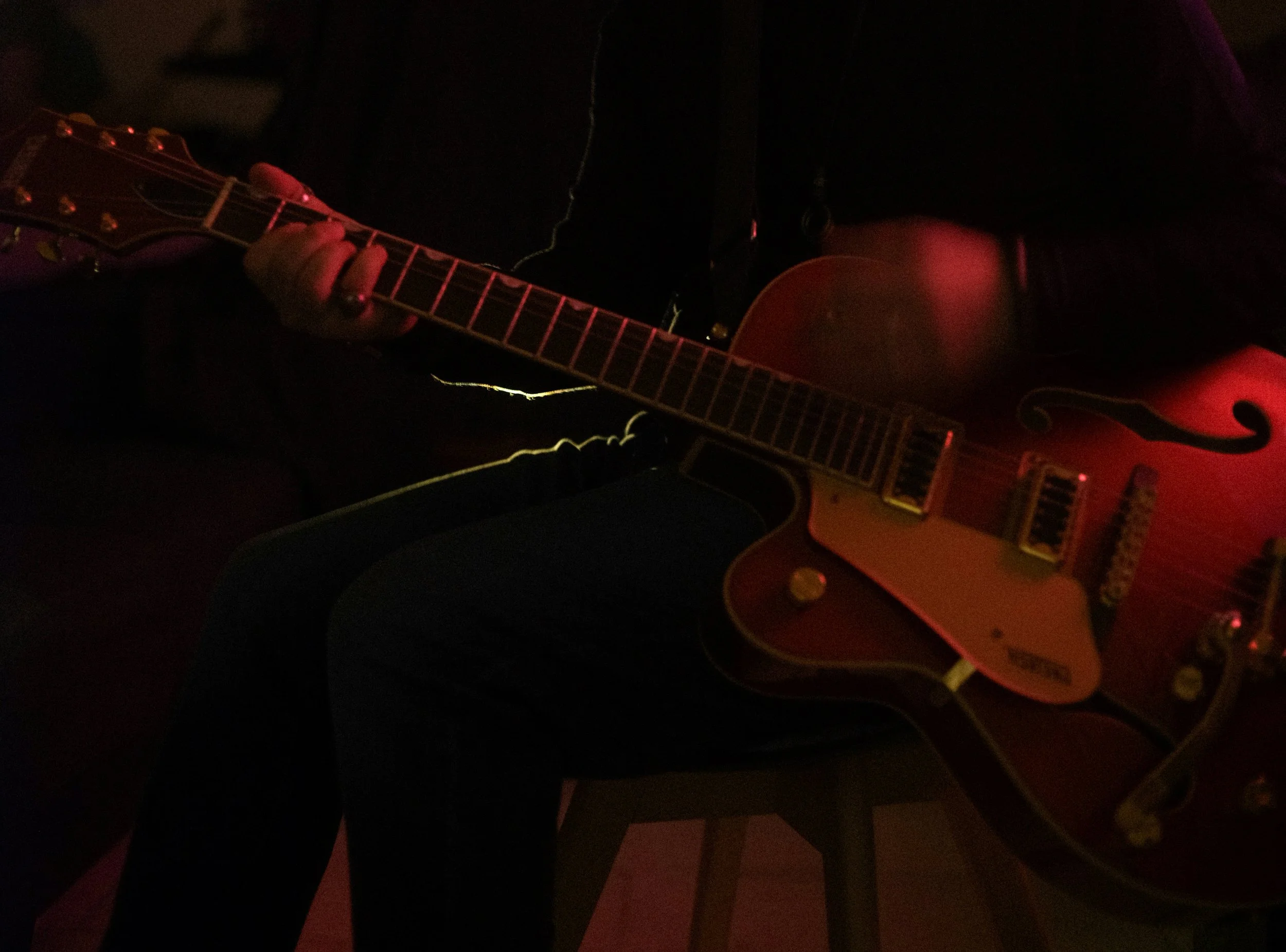 A person is playing an acoustic guitar in a dark setting, illuminated by dim, pinkish-red lighting.