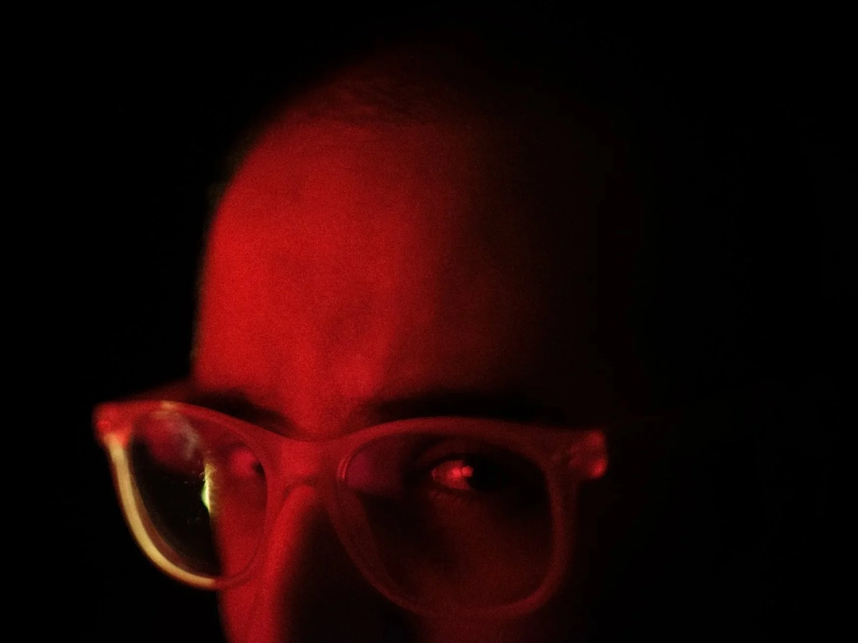 A person wearing glasses with red lighting reflecting off of them, looking down in a dark environment.