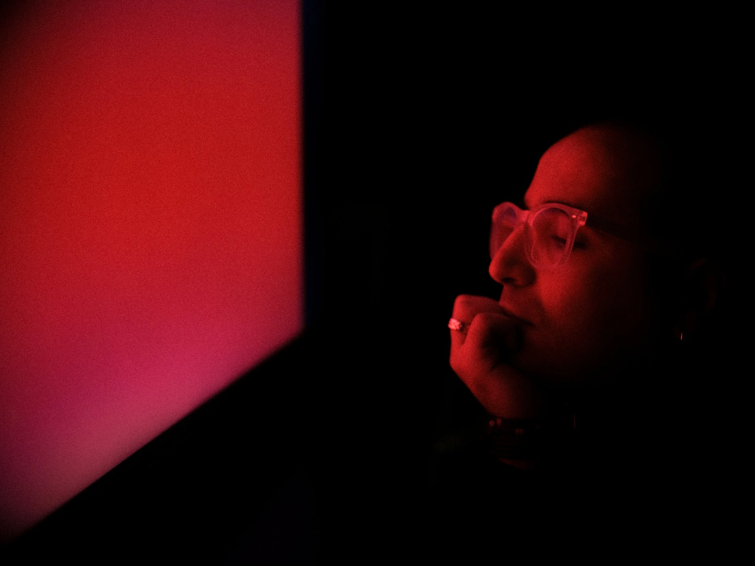 A person wearing glasses is in front of a dark background with a red light source. The person is resting their chin on their hand and appears to be looking at a screen or device.