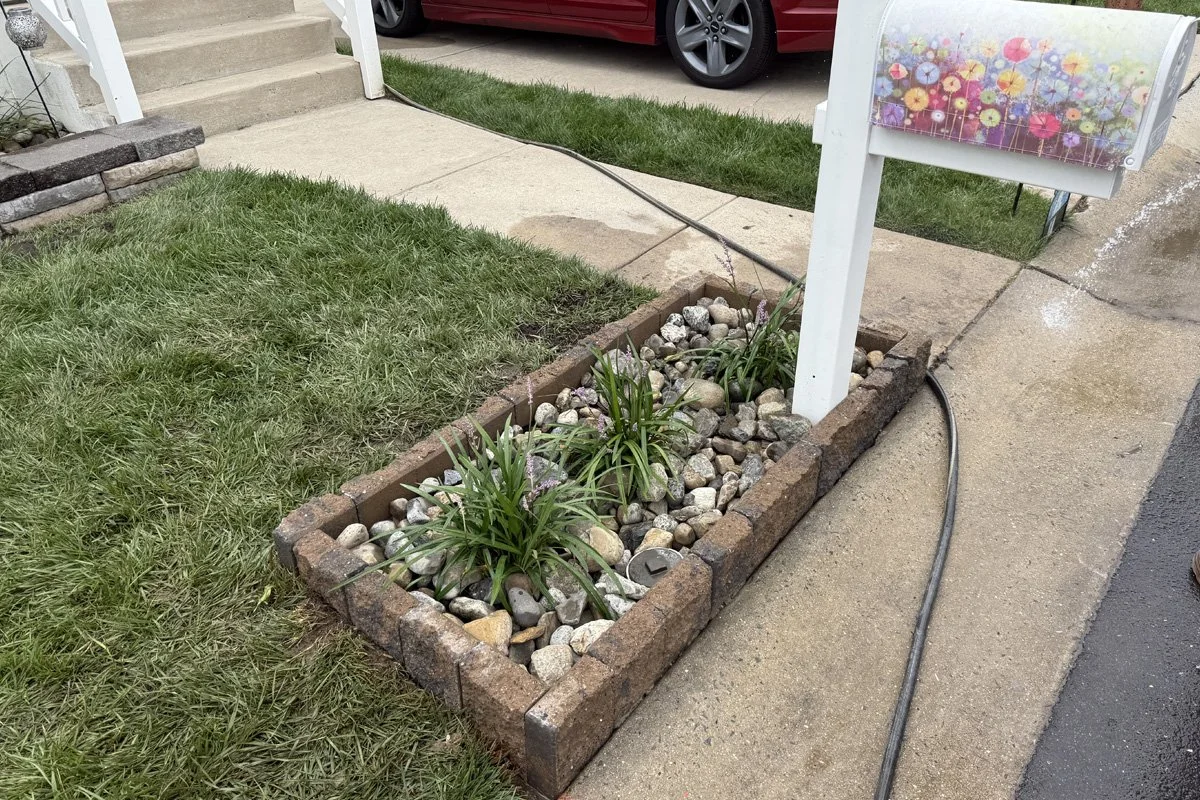 A custom curbside mailbox planter installed by JPL Landscaping in Williamstown, NJ, featuring Belgard Edgestone in Dakota Blend, filled with 1-to-3-inch Delaware river rock and variegated liriope plants.