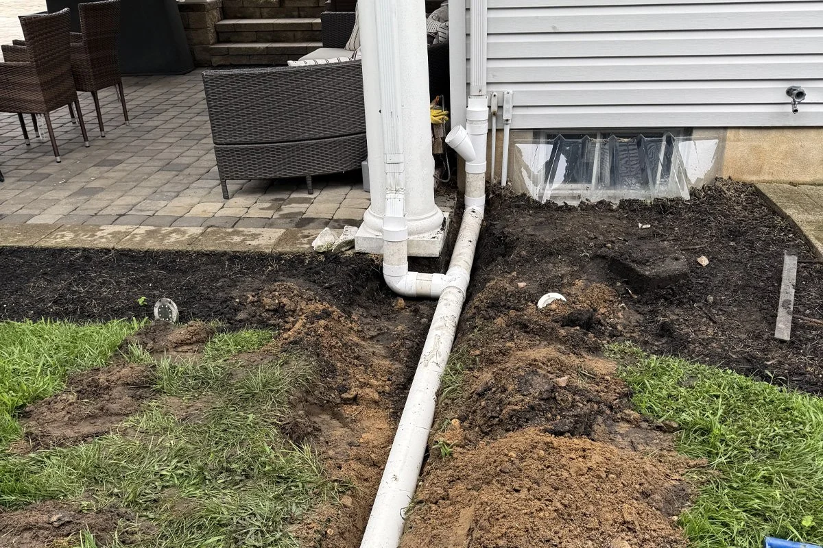 An in-progress underground drainage system by JPL Landscaping in Washington Township, NJ, showing two gutter downspouts tied into a PVC pipe trench terminating at a green NDS pop-up drainage emitter.