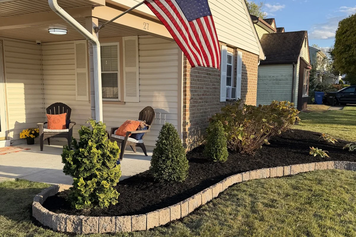 Front porch view of a landscape renovation by JPL Landscaping in Washington Township, NJ, showcasing a curved stone block garden wall, fresh black mulch, and manicured evergreen shrubs under an American flag.