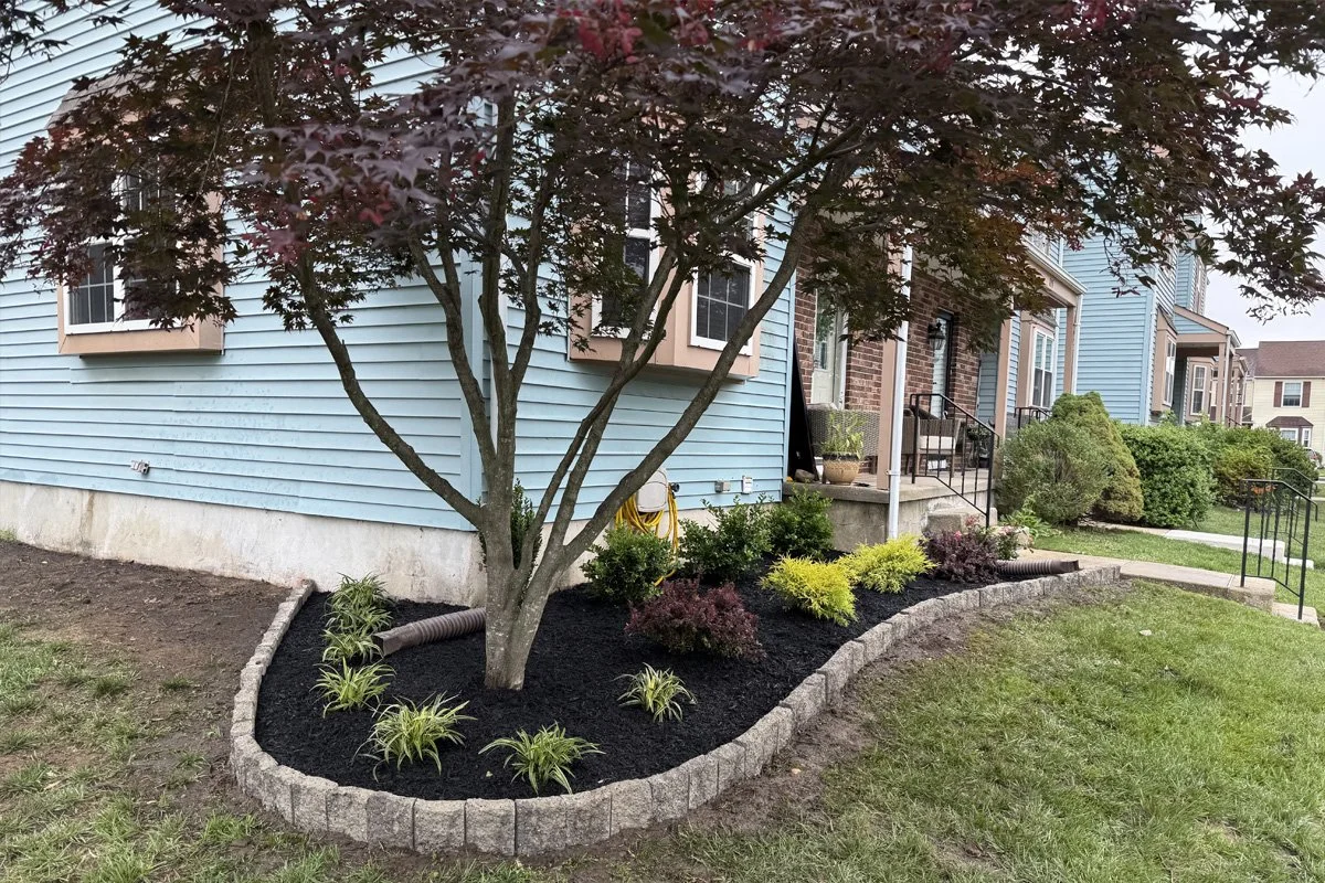 A freshly renovated corner flower bed by JPL Landscaping in West Deptford, NJ, featuring a curved stacked-stone border, rich black mulch, and newly installed ornamental shrubs.