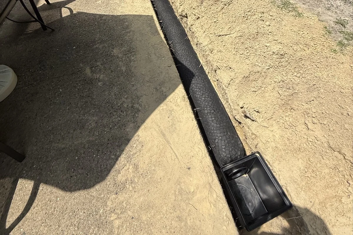Installation of a heavy-duty NDS catch basin box and a fabric-wrapped drainage pipe system in a dirt trench alongside a concrete patio.