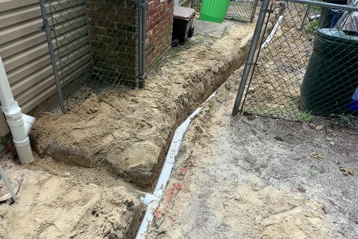 A long outdoor drainage trench containing a white PVC stormwater pipe run with a Y-connector passing a brick chimney and chain-link fence.