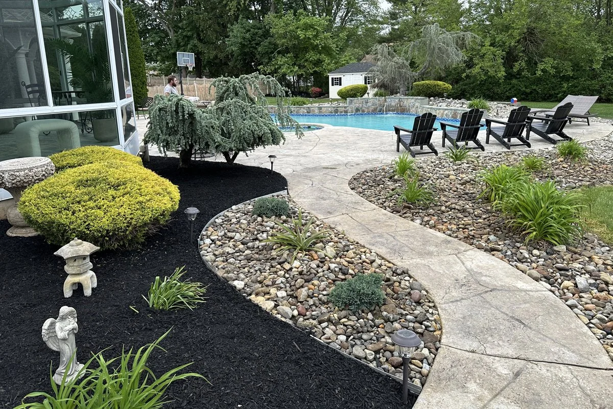 A wide-angle look at a premium backyard swimming pool area landscaped by JPL Landscaping, featuring black lounge chairs, stone walkways, and vibrant garden beds.
