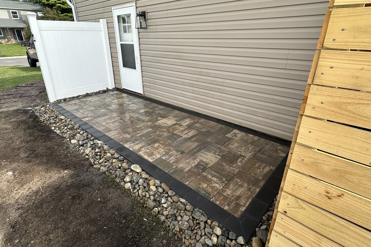 A newly installed multi-colored interlocking paver patio pad outside a back door, featuring a clean dark border and a decorative river rock drainage edge.