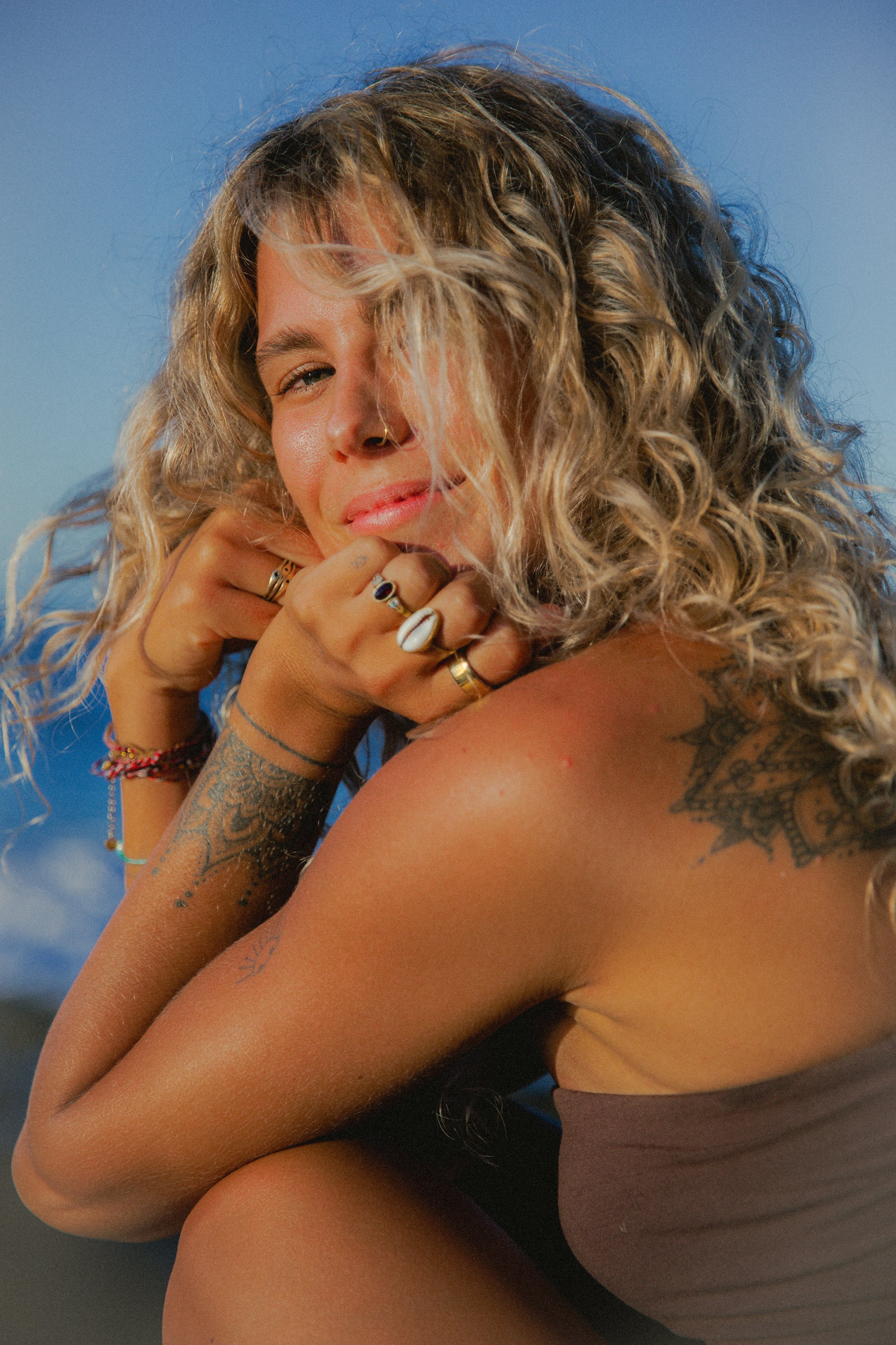 A woman with curly blonde hair and tattoos on her arm, wearing rings, posing in front of a blue sky with clouds.