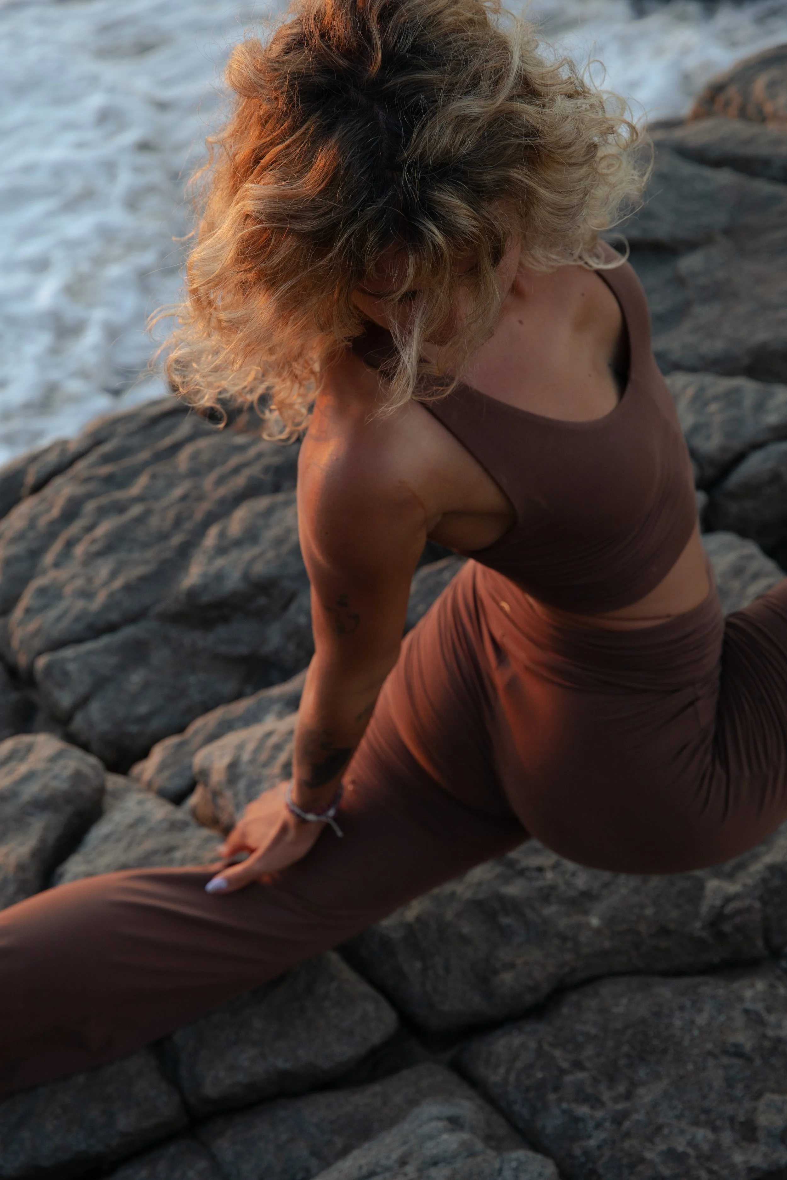 A woman with curly blonde hair sitting on rocks near water, wearing a brown tank top and pants, looking down and to the side.