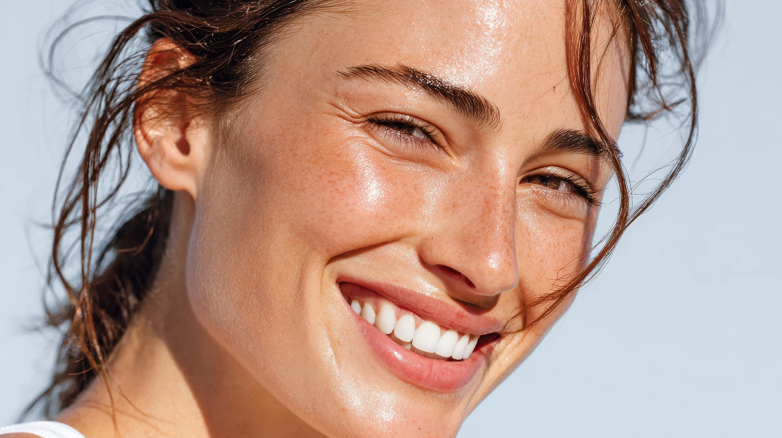 Close-up of a smiling woman with sparkling skin and wet hair, outdoors with a bright sky background.
