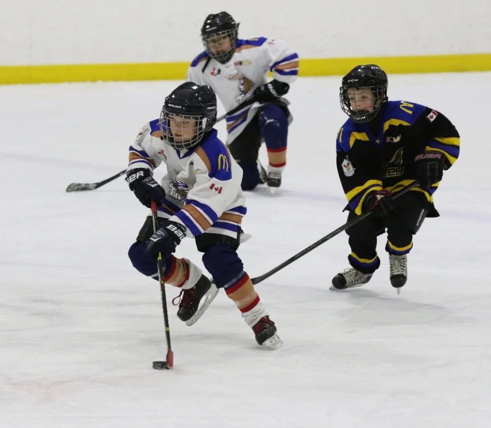 Three young children playing hockey on an ice rink, wearing helmets and jerseys.