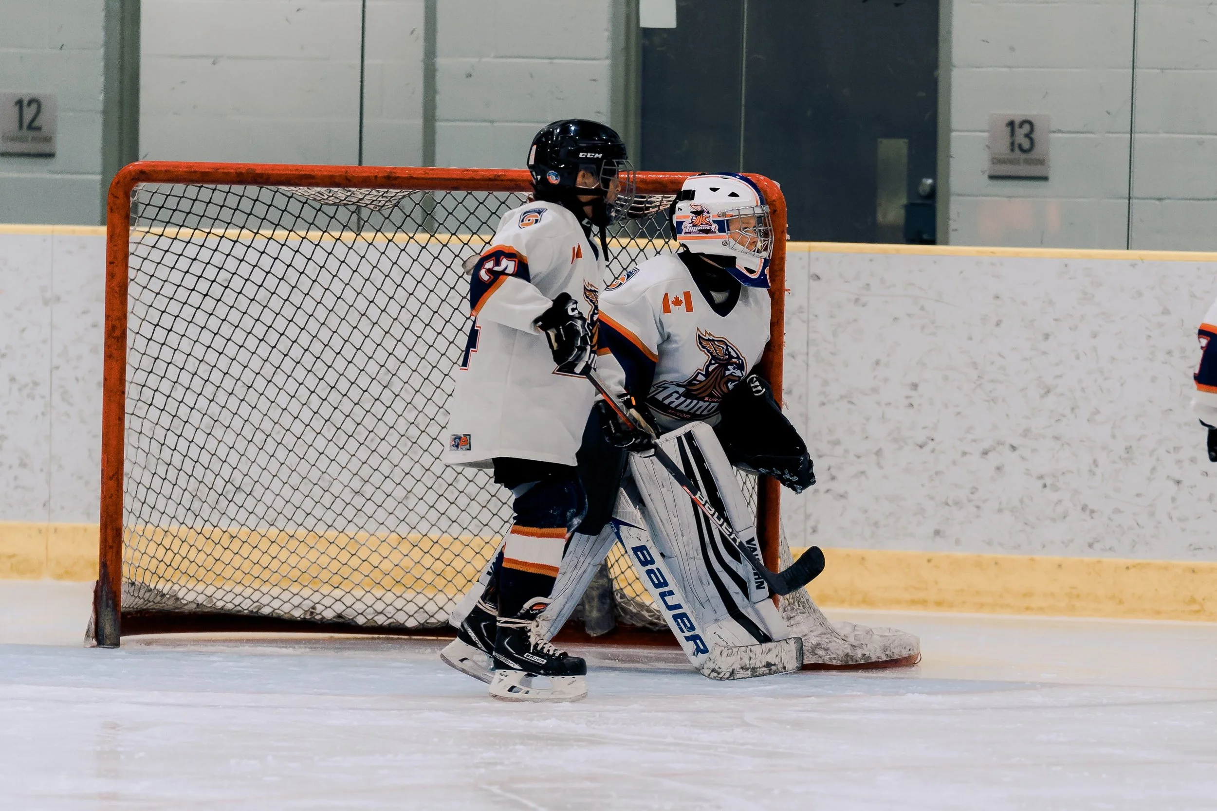 An ice hockey goalie standing in front of the goal net on the ice rink, wearing a white uniform with team logos and protective gear, holding a hockey stick, with another player partially visible on the right side.