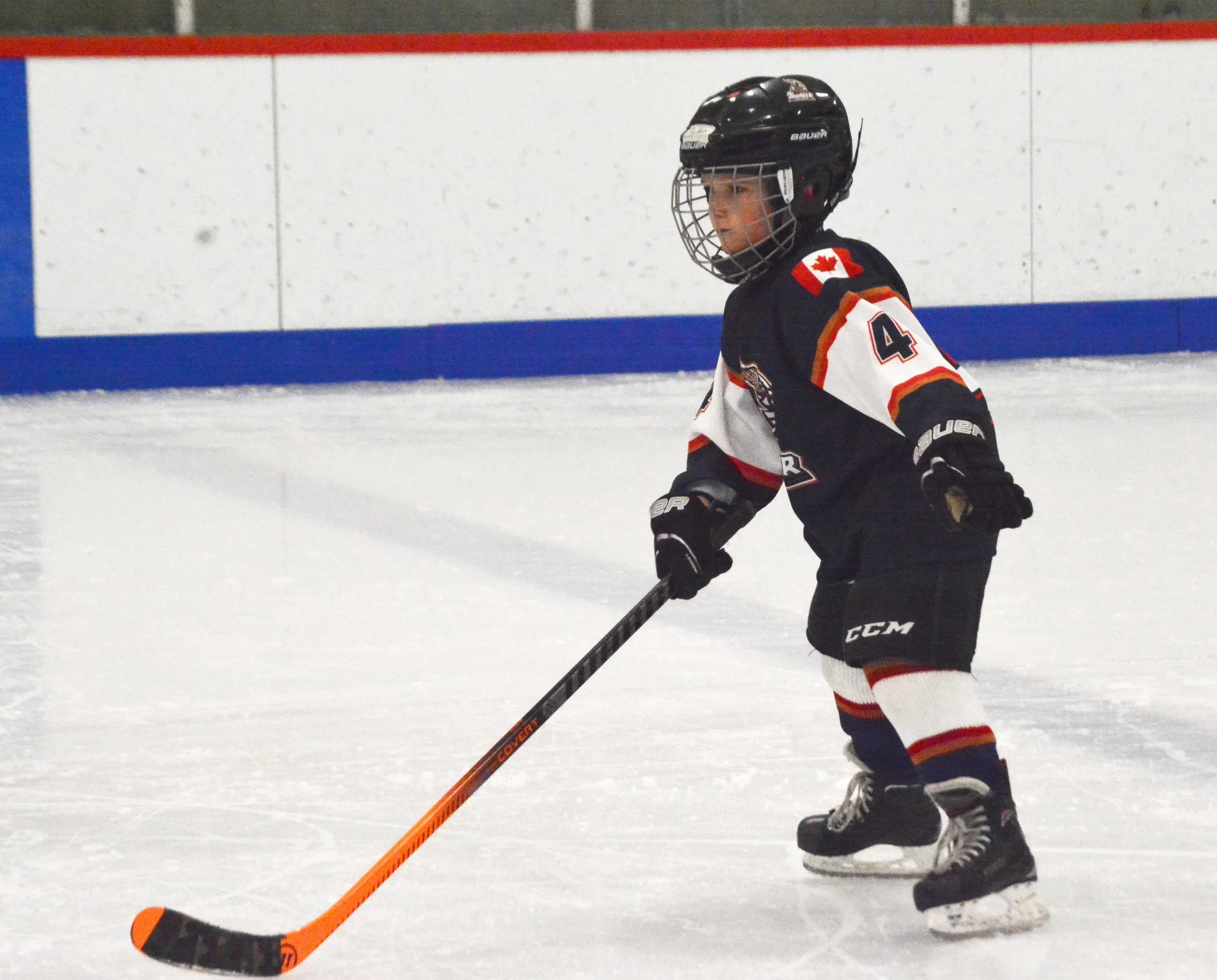 A young hockey player in black, white, and red team uniform, wearing a helmet with a Canadian flag patch, playing on an ice rink with a red, white, and blue barrier in the background.