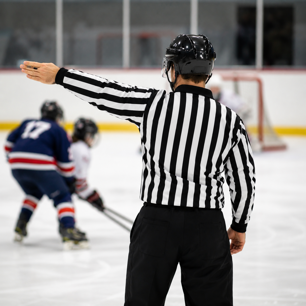 Referee in black and white striped shirt and black helmet signaling during an ice hockey game, with players in the background near the goal.