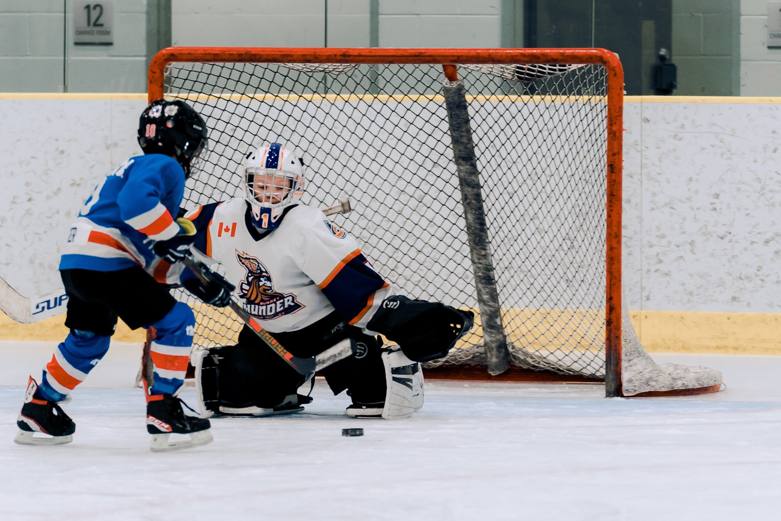 Ice hockey game with a young goalie in a white jersey and a player in a blue jersey near the net on an indoor rink.
