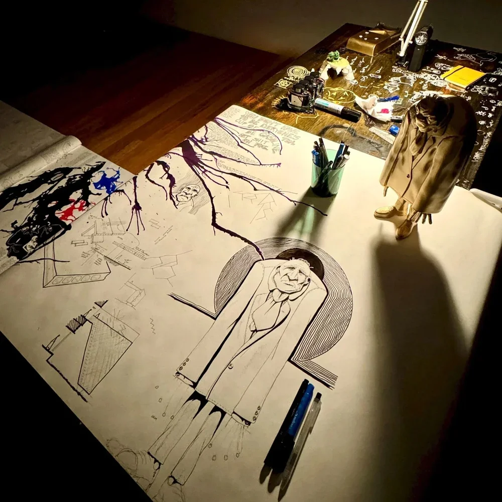 A table with various art supplies, including pens, markers, and notebooks. Several sketches and drawings are laid out, featuring a caricature of a person in a coat, a tree with roots and branches, and other abstract designs.