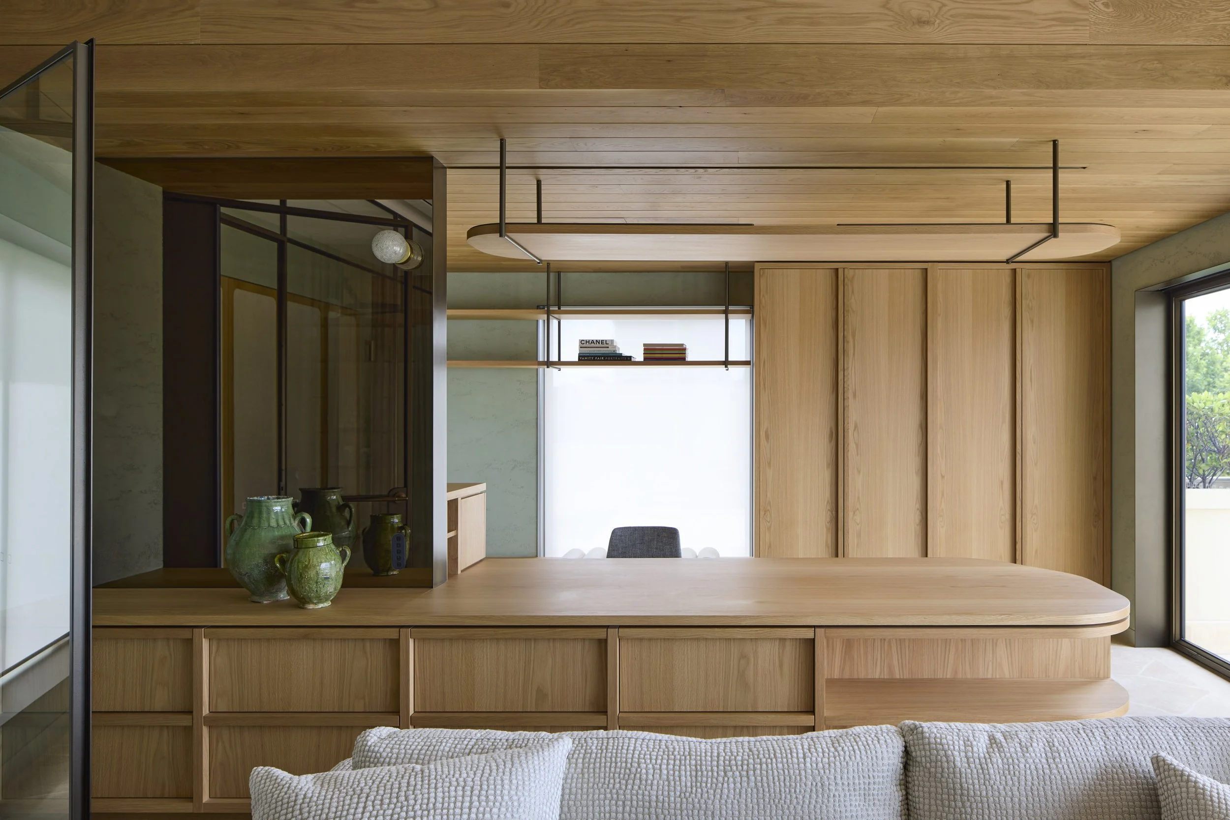 Interior of a modern home with wooden ceiling, wall paneling, and a built-in bookshelf with books. \Green pottery decor is on a wooden ledge, and part of a beige sofa is visible in the foreground.